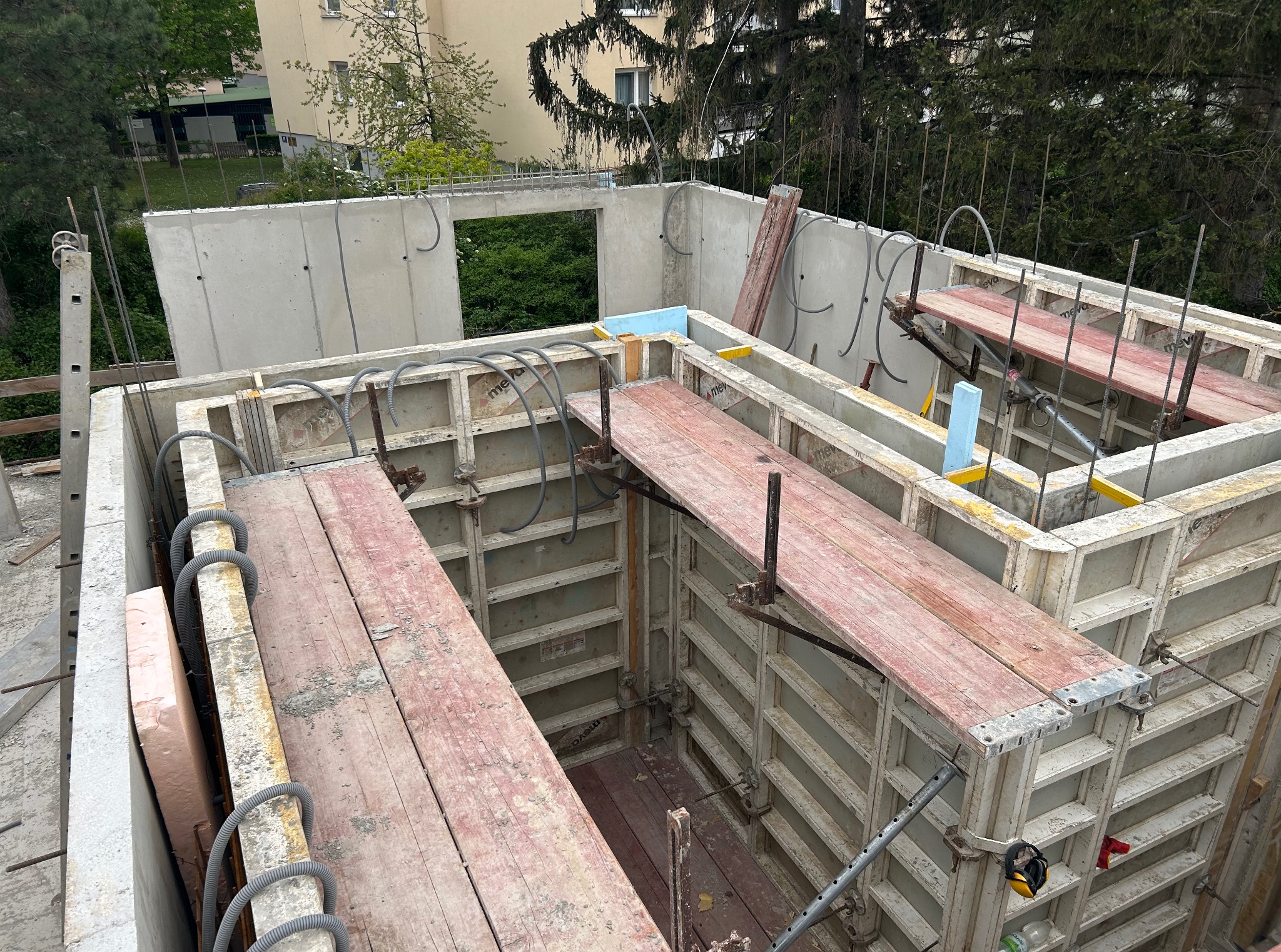 Szare szalunki fundamentów z deskami pomostowymi, widok z góry na budowę, z zielenią w tle i elementami instalacji. W tle budynek mieszkalny.