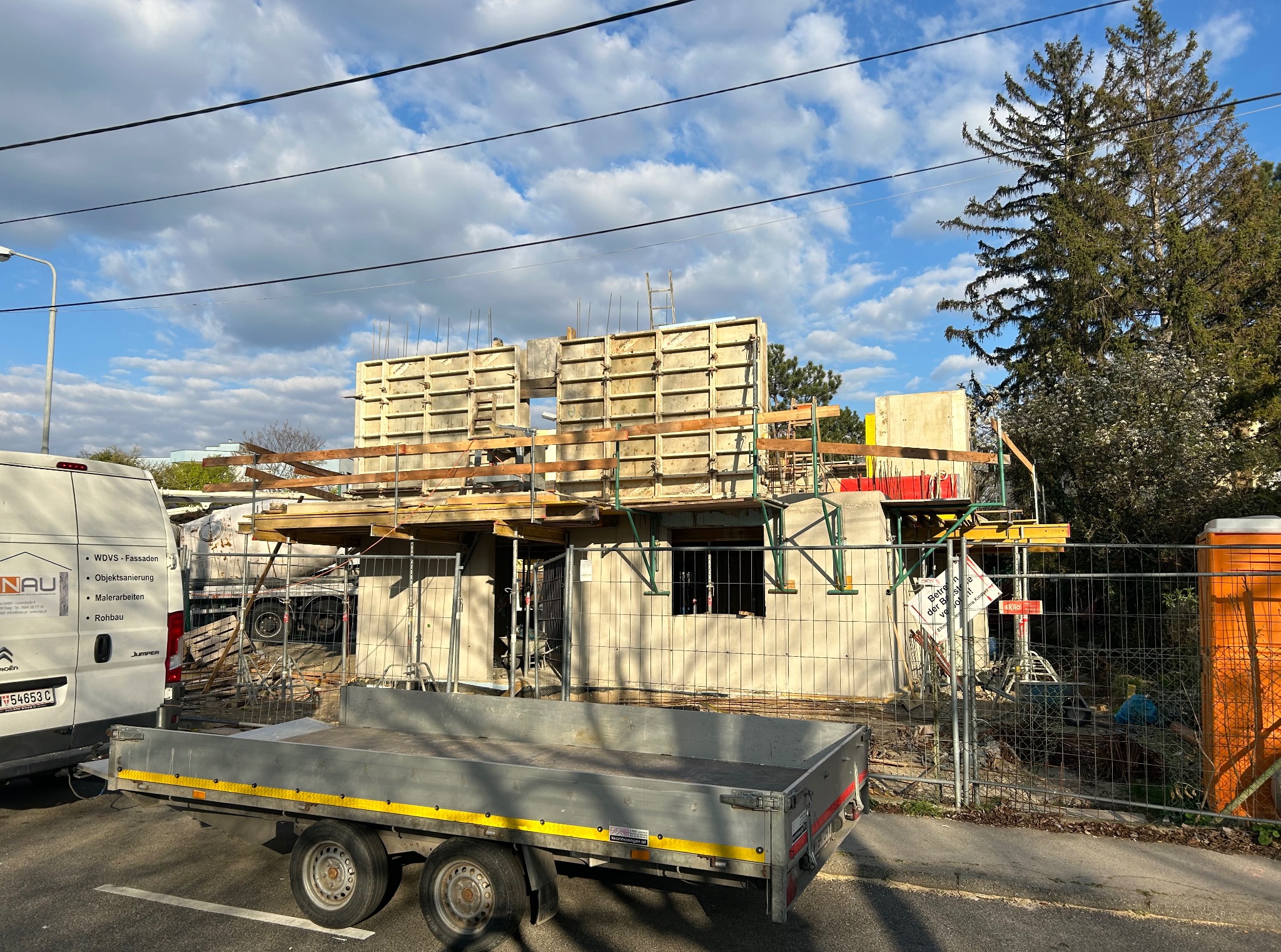 Wznoszona ściana domu z betonu komórkowego, widoczne szalunki i zbrojenia. Na pierwszym planie przyczepka i częściowo widoczny samochód dostawczy.