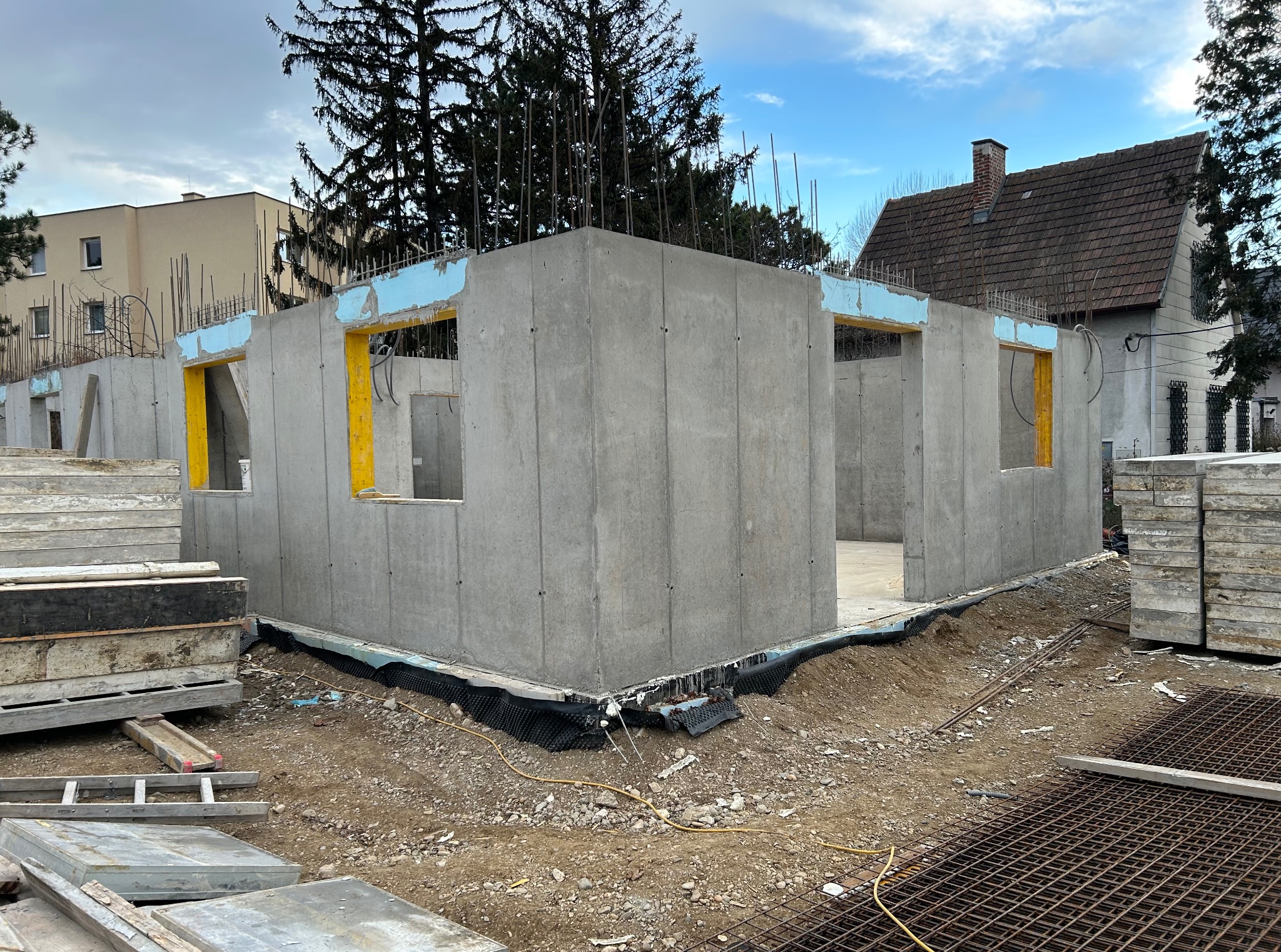 Surowy stan ścian parteru domu z betonu komórkowego, widoczne żółte ramy okienne oraz wystające pręty zbrojeniowe, teren budowy z elementami konstrukcyjnymi.