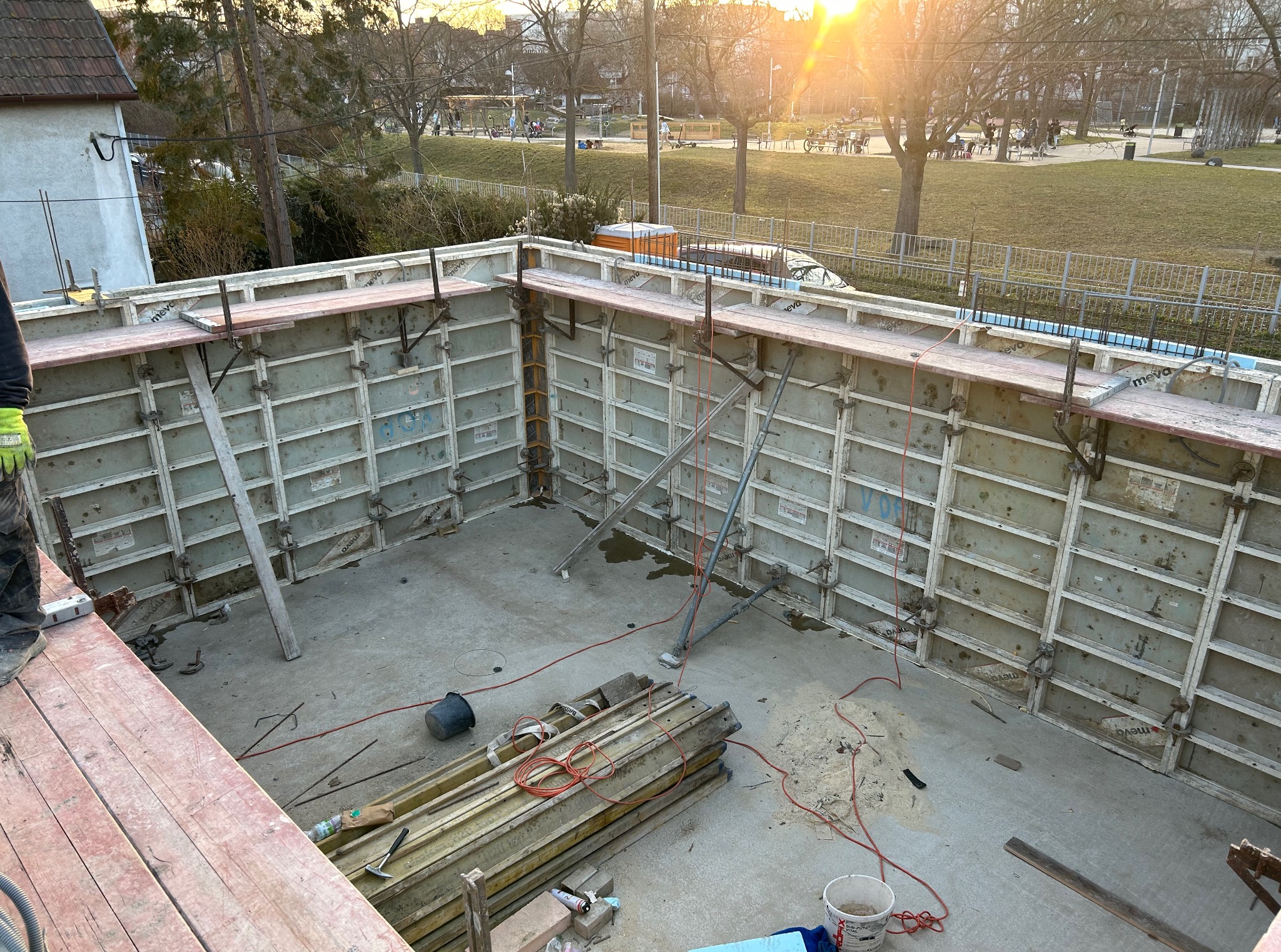Zalany fundament z deskowaniem MEVA, widok z góry. Na pierwszym planie deski, wiadro i narzędzia. W tle park miejski w promieniach zachodzącego słońca.