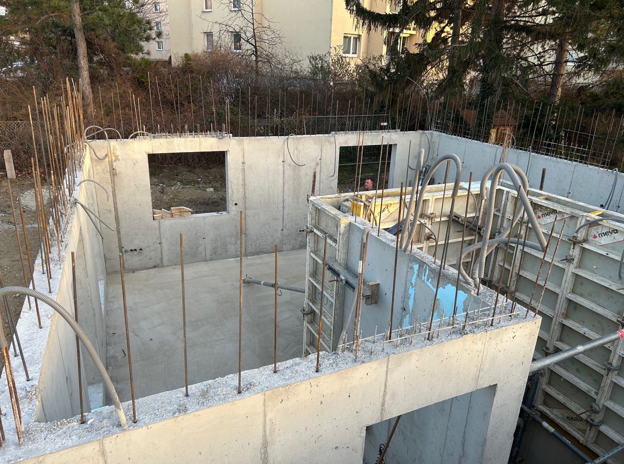 Wylane fundamenty z otworami okiennymi, wystającymi prętami zbrojeniowymi. Widoczne szalunki i peszle instalacyjne. W tle budynki mieszkalne i drzewa.