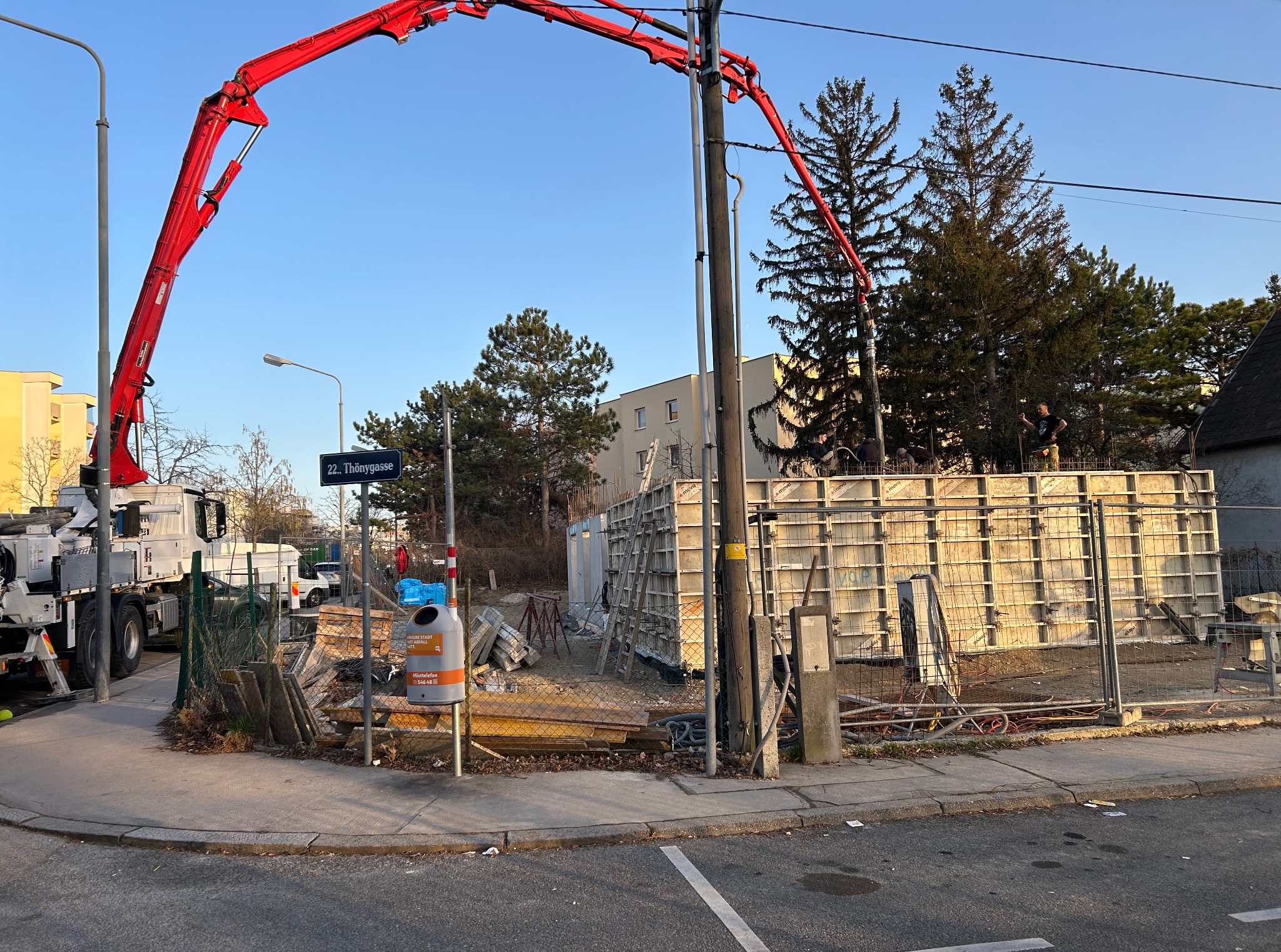 Wylewanie fundamentów: czerwony wysięgnik pompy do betonu nad szalunkiem, widoczni robotnicy. Ulica '22. Thönygasse' w tle. Słoneczny dzień.