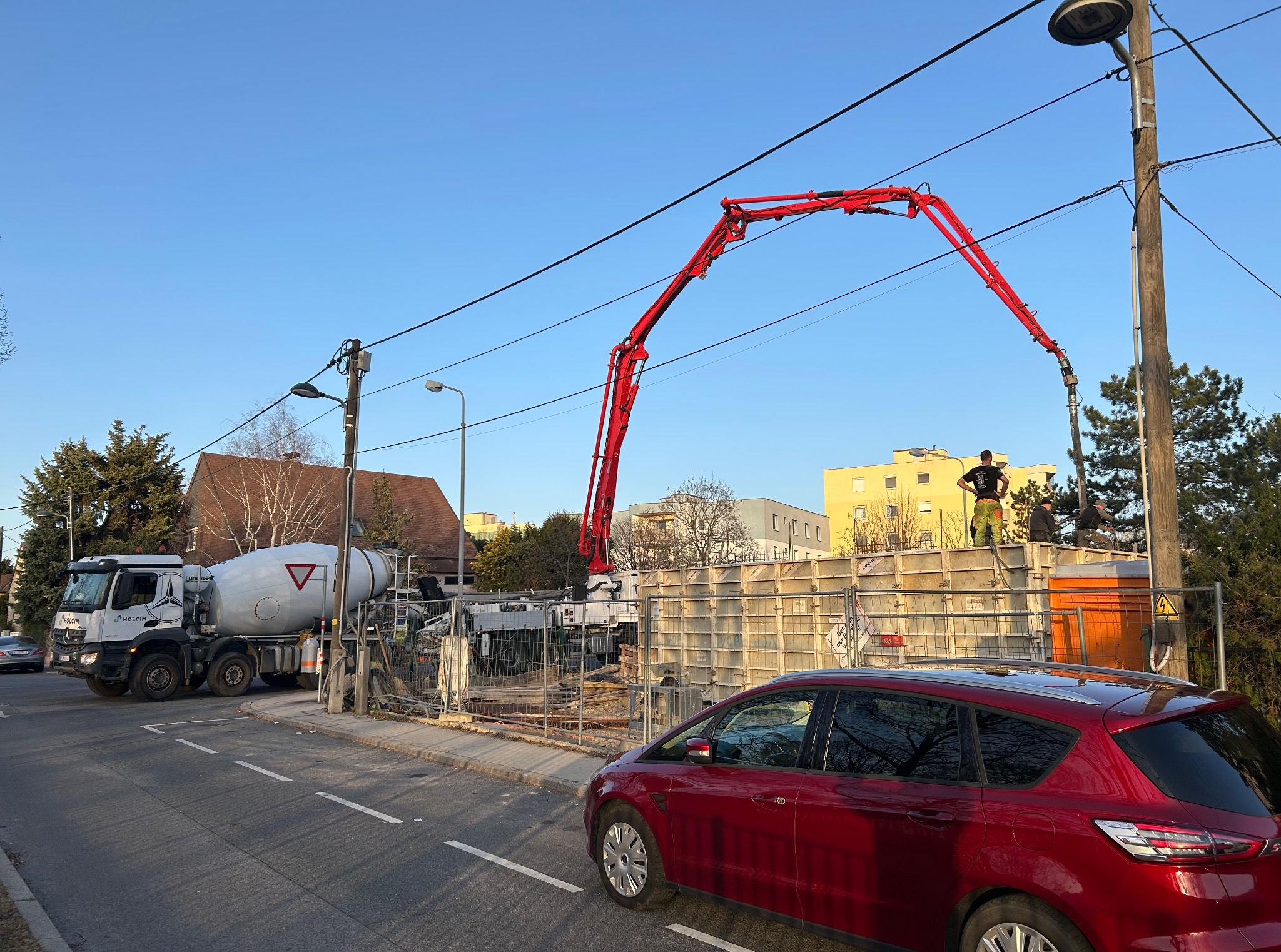 Wylewka fundamentów: betonowóz Holcim, pompa do betonu i pracownicy na szalunku. Czerwony samochód osobowy na pierwszym planie.