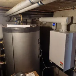 Nowoczesny piec gazowy Viessmann zamontowany w piwnicy, z podłączonym zasobnikiem CWU i orurowaniem. Widoczne elementy instalacji i detektor gazu.