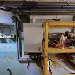 Metgen Service Krzysztof Rekus - Biały piec gazowy Viessmann zamontowany na niebieskiej, częściowo wykończonej ścianie w piwnicy, z rurami i półką z narzędziami obok.