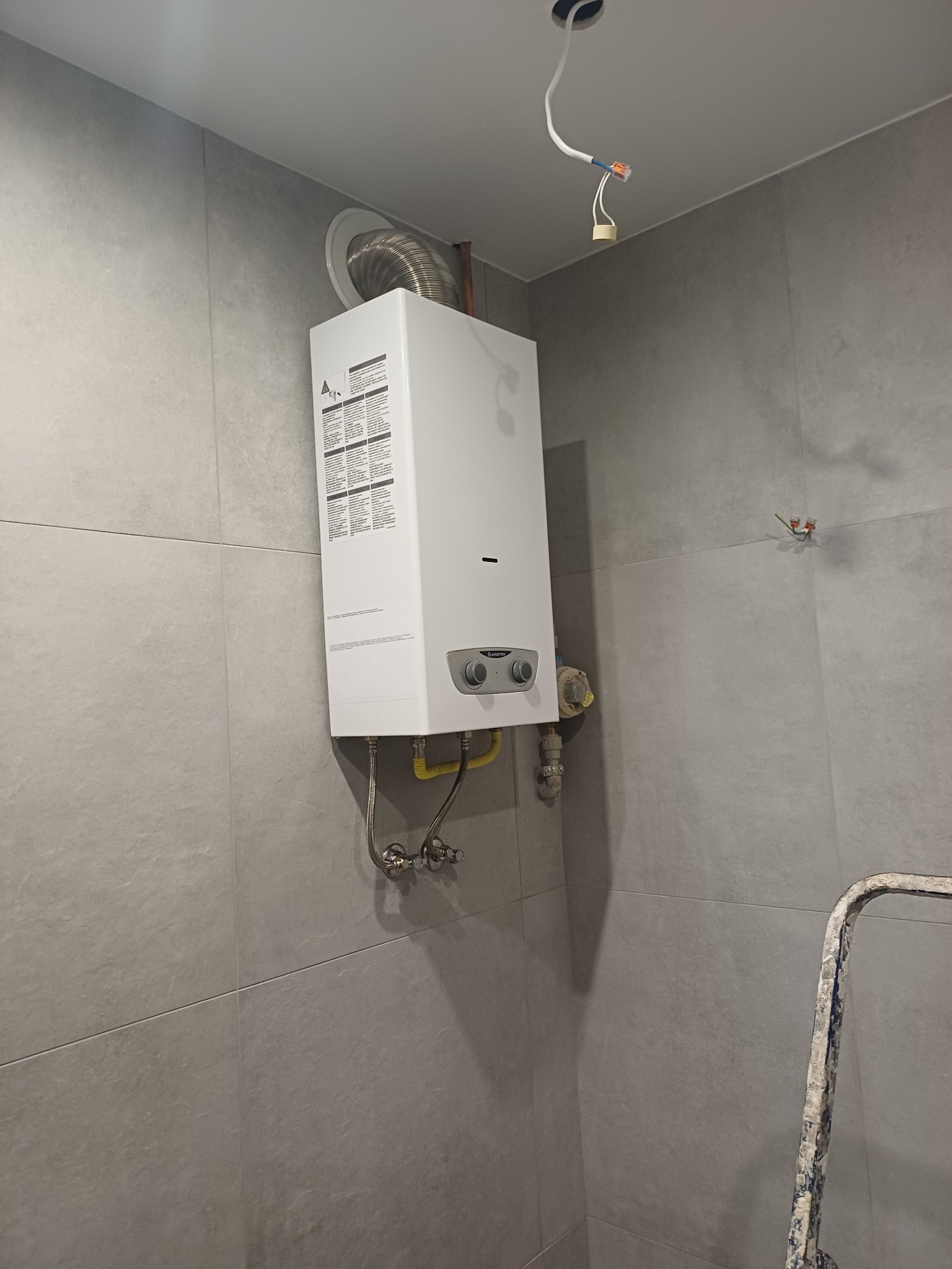 Biały piec gazowy Ariston zamontowany na szarej, kafelkowanej ścianie. Widoczne rury doprowadzające gaz i woda oraz przewód kominowy. W tle drabina malarska.