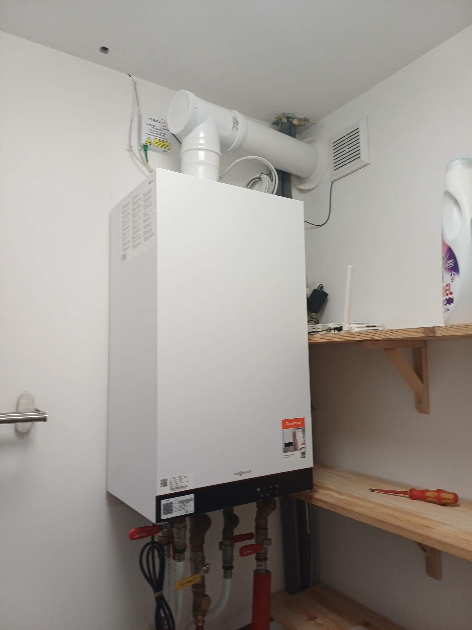 Biały piec gazowy Viessmann zamontowany na ścianie w pomieszczeniu gospodarczym, z widocznymi rurami i elementami instalacji. Na półce leży czerwony śrubokręt.