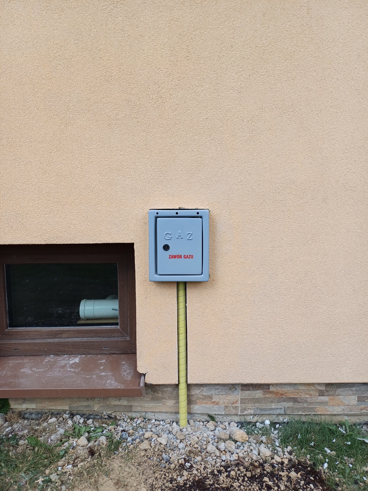 Szara skrzynka gazowa z czerwonym napisem 'Zawór gazu' na beżowej elewacji budynku, obok okna piwnicznego i żółtej rury. U dołu widoczne kamienie i trawa.