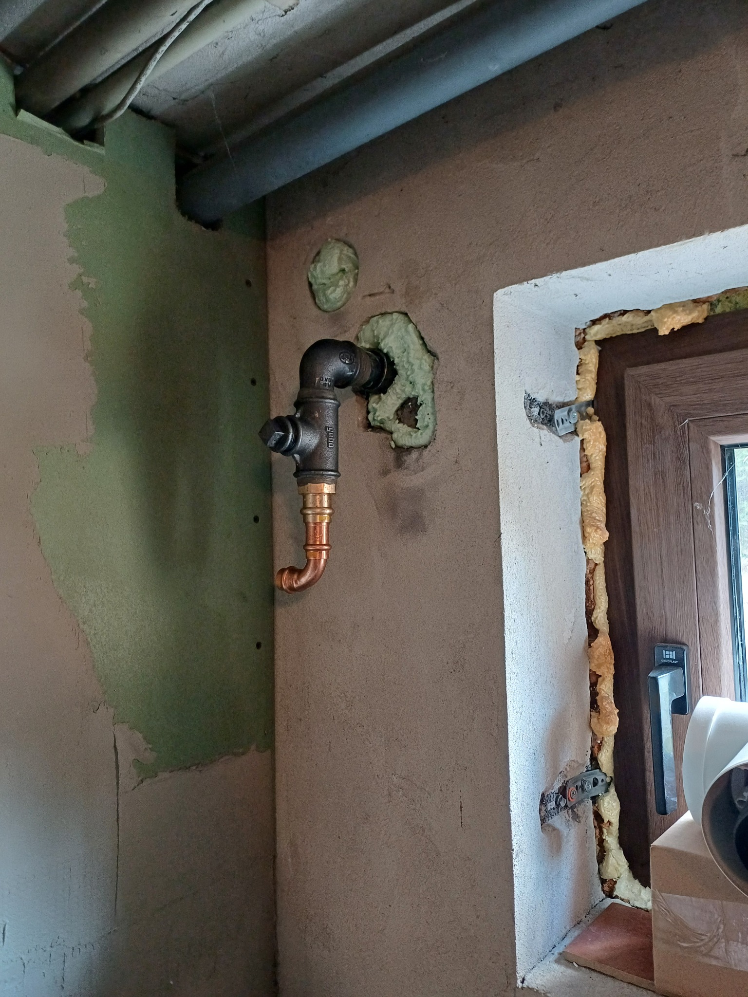 Fragment ściany z zamontowaną, miedziano-stalową instalacją hydrauliczną, zabezpieczoną pianką montażową przy oknie. Widoczne niedokończone prace tynkarskie.