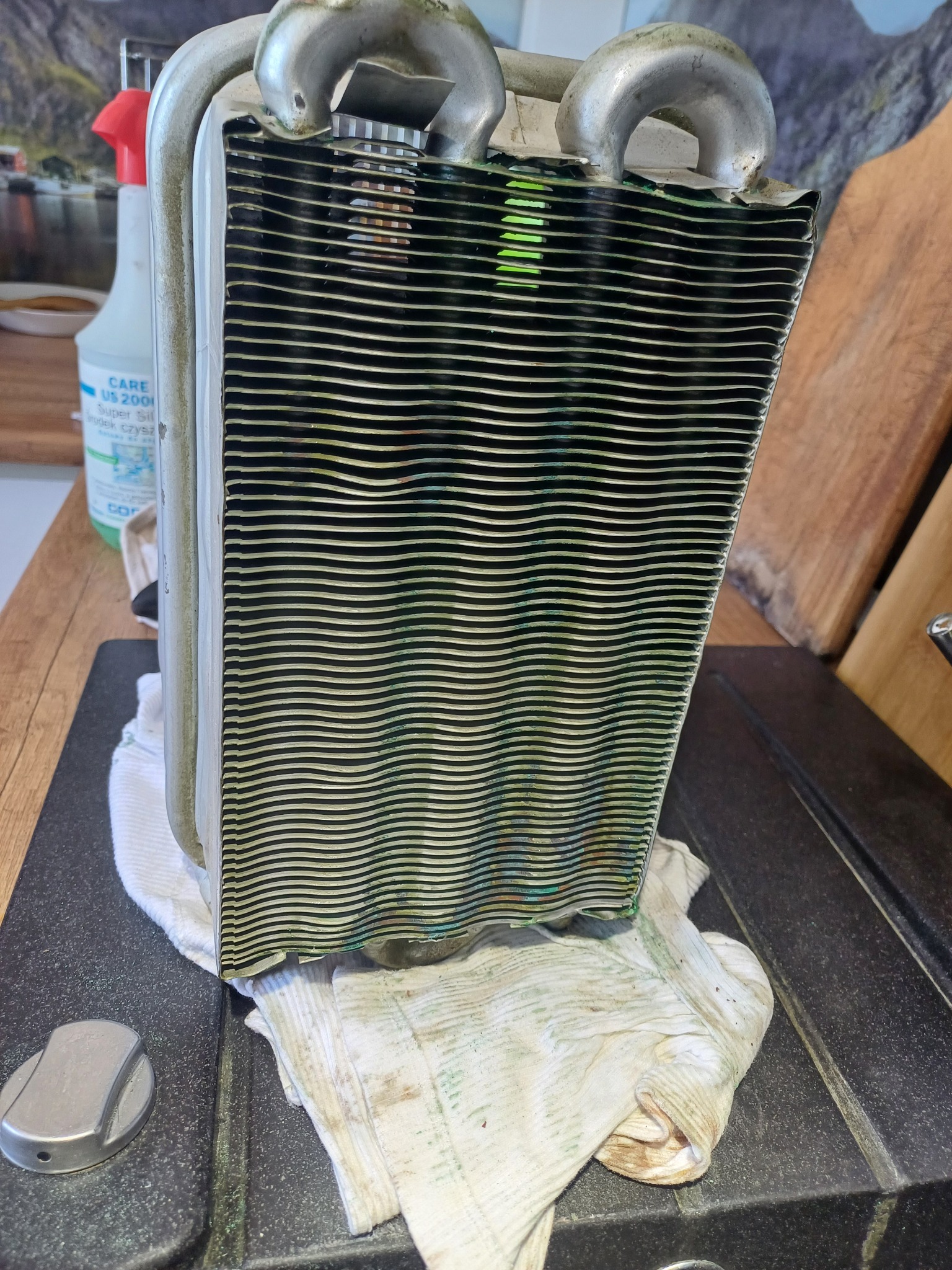 Wymiennik ciepła z pieca gazowego, widoczne ślady użytkowania i czyszczenia. Element leży na ściereczce, w tle butelka środka czyszczącego i fragment blatu.