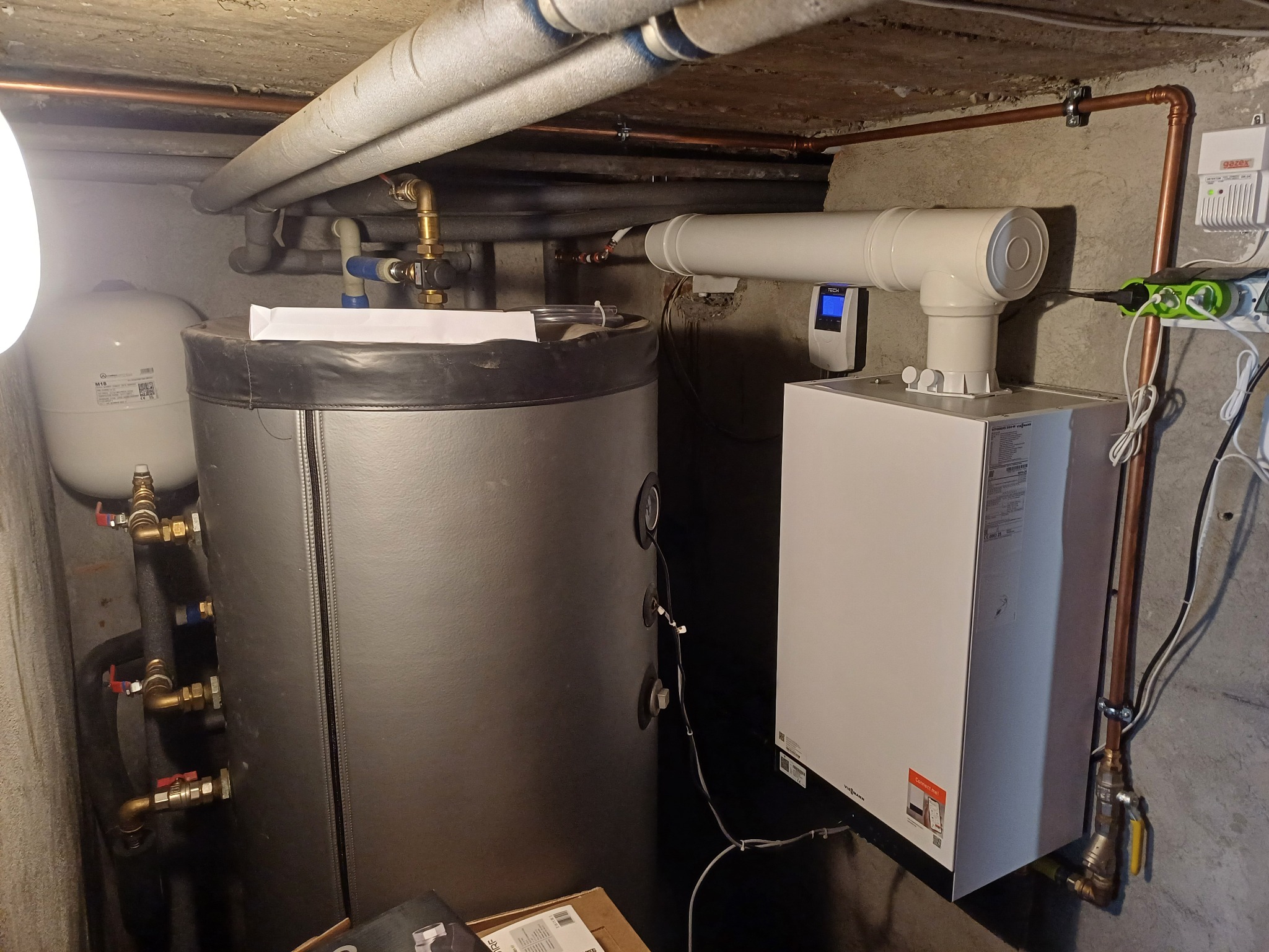 Nowoczesny piec gazowy Viessmann zamontowany w piwnicy, z podłączonym zasobnikiem CWU i orurowaniem. Widoczne elementy instalacji i detektor gazu.