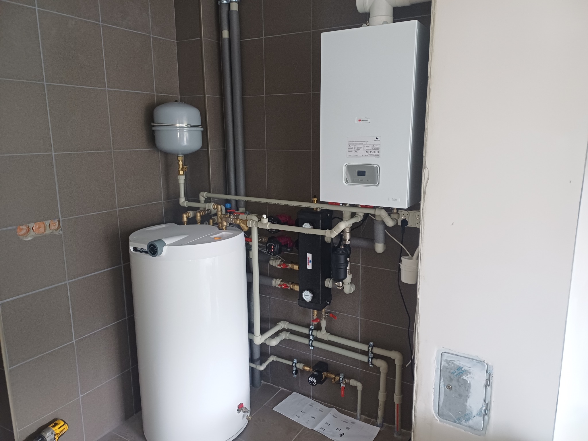 Nowoczesny, biały piec gazowy Viessmann z podłączonym zasobnikiem wody w kotłowni z brązowymi płytkami. Widoczne rury i osprzęt instalacji CO.