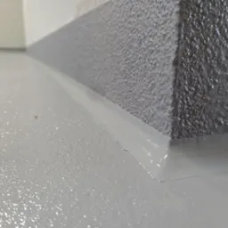 Jasnoszara, gładka posadzka żywiczna w rogu pomieszczenia, z widocznym połączeniem ściany i podłogi. Delikatna faktura ściany. Minimalistyczny detal wykończenia.