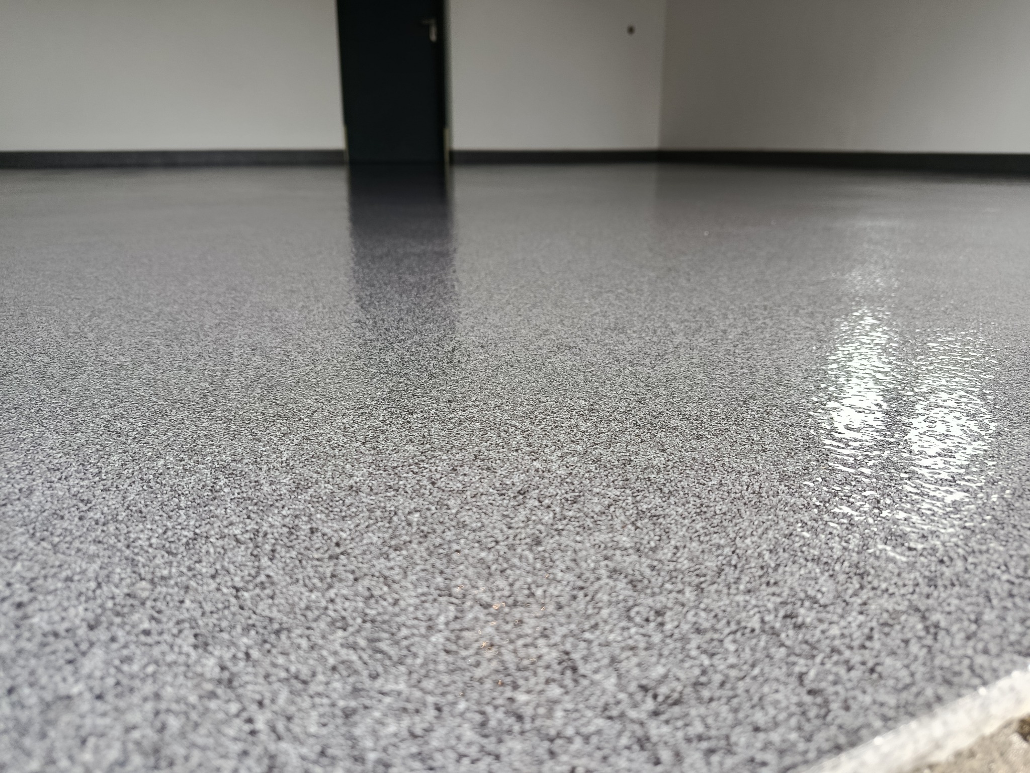 Błyszcząca, szara posadzka żywiczna z drobnym wzorem w pustym pomieszczeniu. Widoczne odbicie światła na powierzchni, czarne listwy przypodłogowe i drzwi w tle.