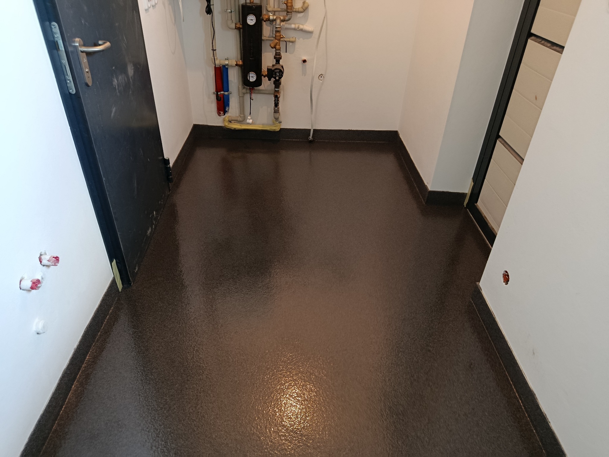 Błyszcząca, brązowa posadzka żywiczna w małym pomieszczeniu z instalacją grzewczą i dwiema parami drzwi. Widoczny detal wykończenia przy ścianach.