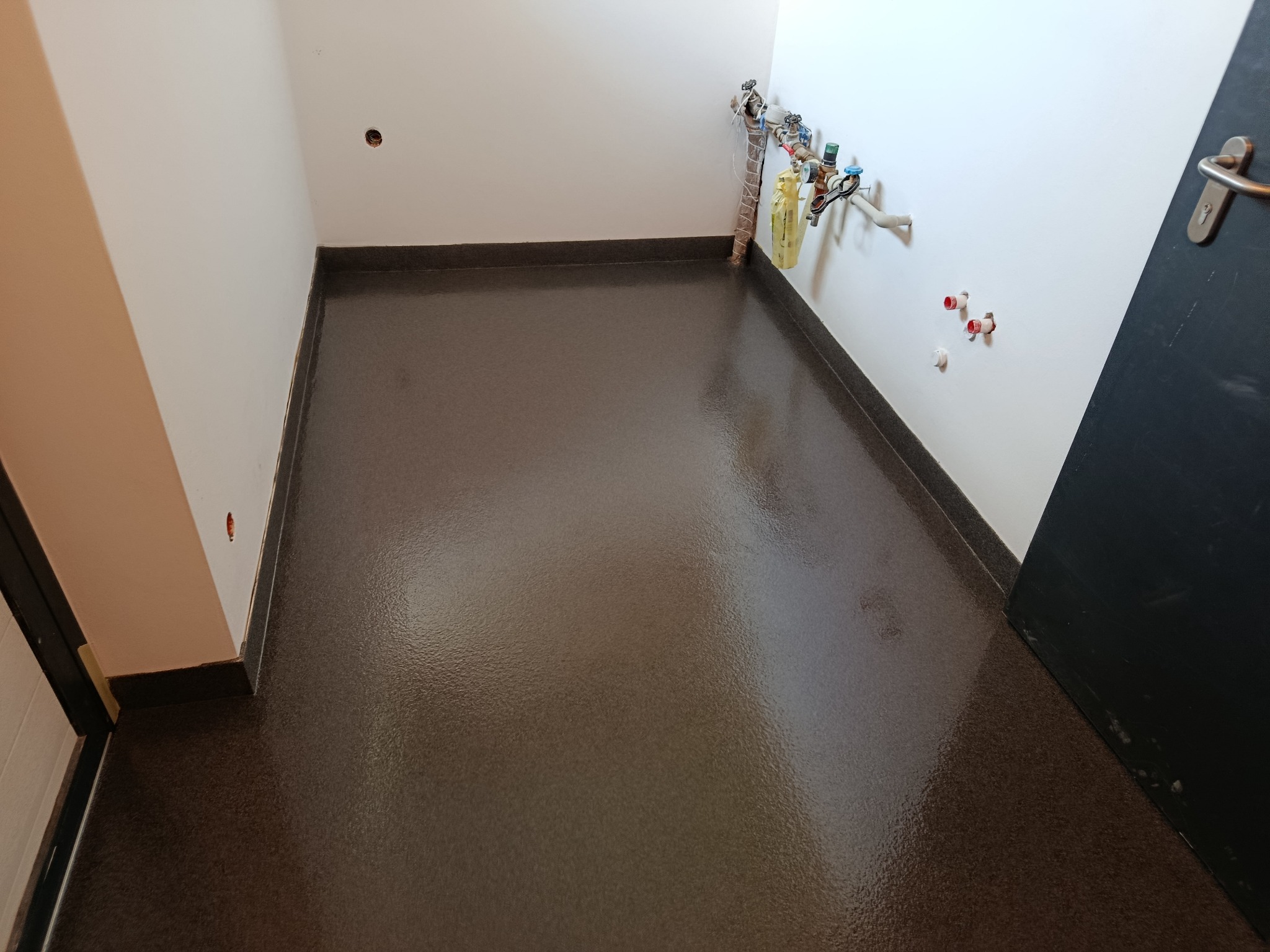 Gładka, brązowa posadzka żywiczna w pomieszczeniu z białymi ścianami i widocznymi instalacjami hydraulicznymi. Ciemne listwy przypodłogowe, drzwi po prawej.