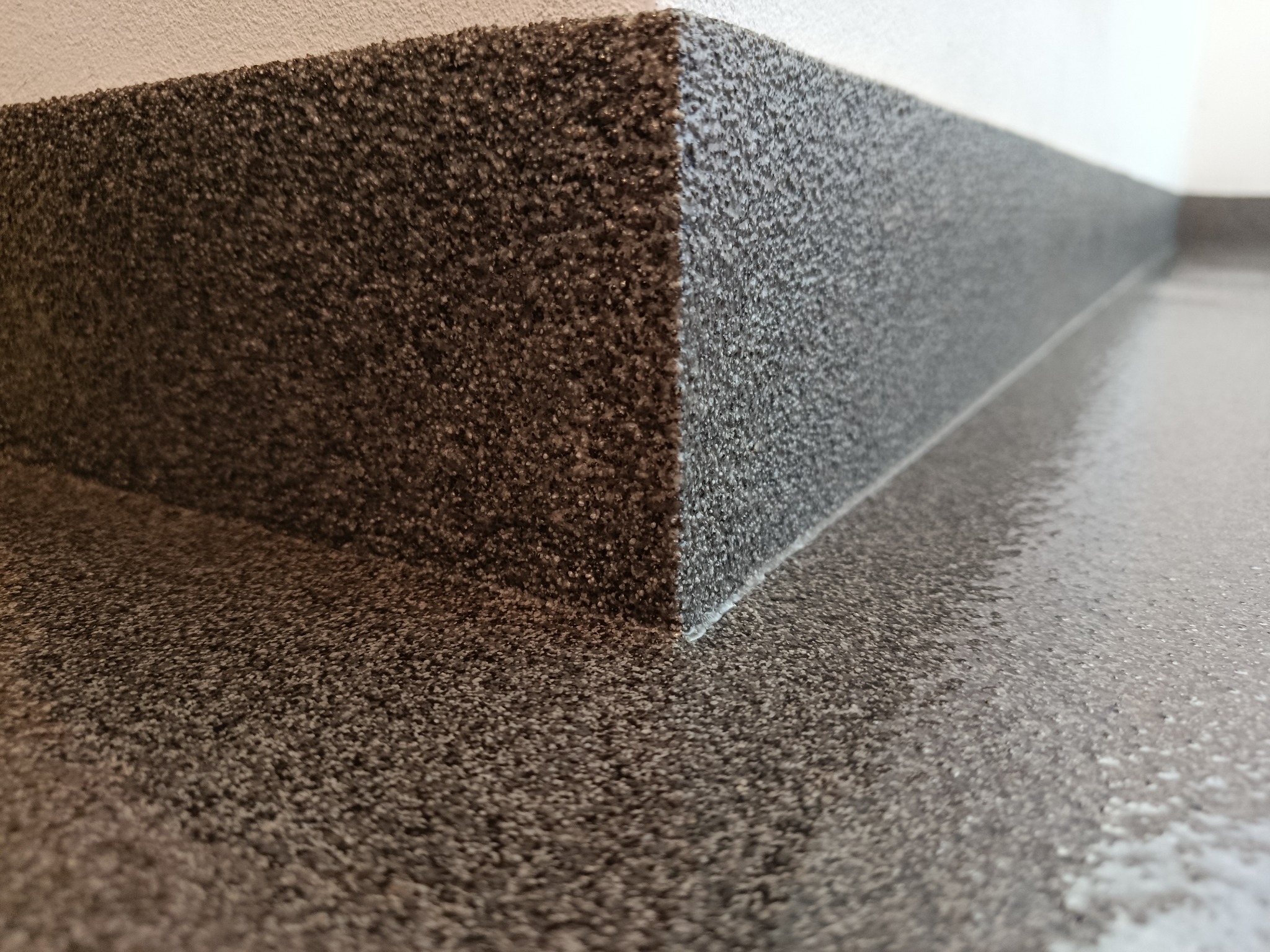 Szara, błyszcząca posadzka żywiczna z wykończoną listwą przypodłogową, zbliżenie na narożnik. Widoczne drobne ziarna w strukturze żywicy.