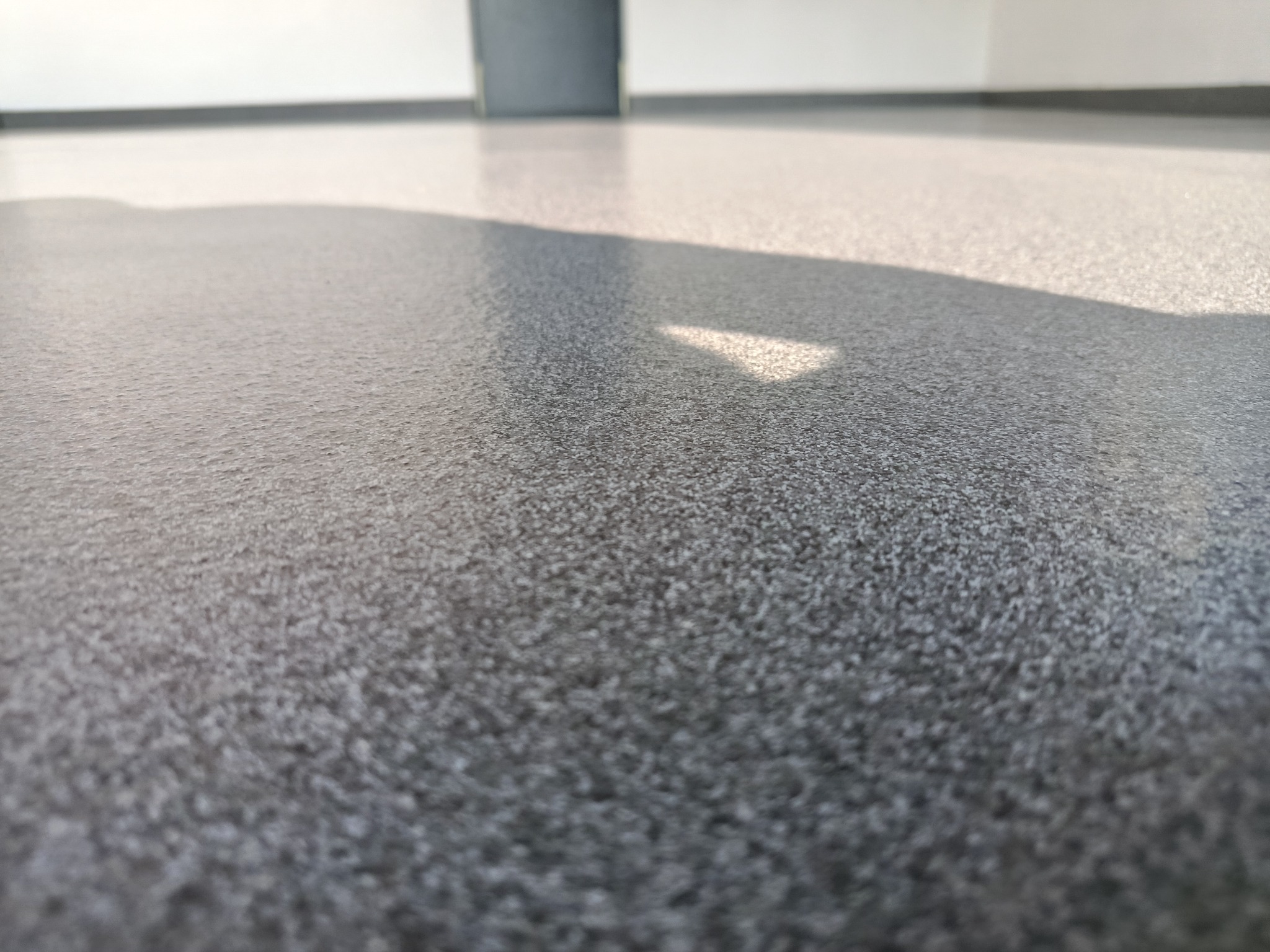 Szara, gładka posadzka z delikatną fakturą, oświetlona naturalnym światłem padającym z okna. W tle widoczne drzwi i fragment ściany.