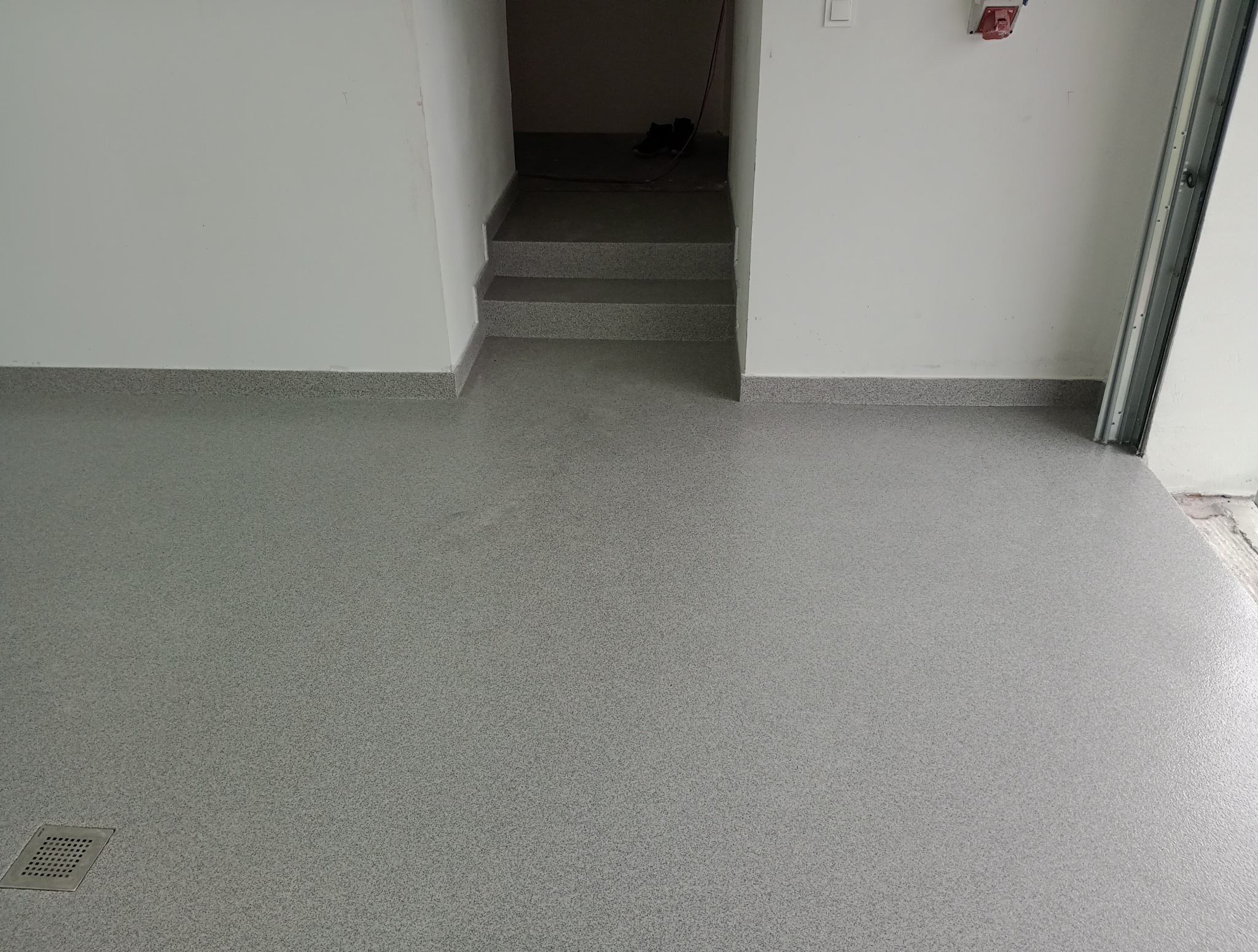 Jasnoszara posadzka żywiczna w garażu z odpływem liniowym, widoczne schody prowadzące w dół i fragment bramy garażowej. Gładka, jednolita powierzchnia.