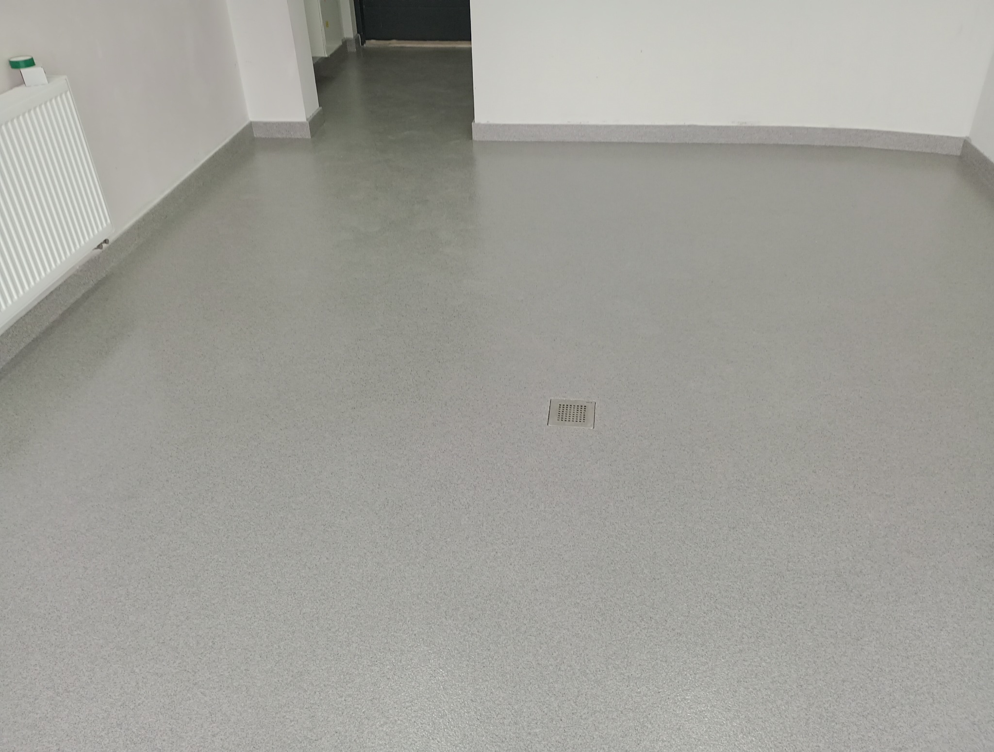 Jasnoszara posadzka żywiczna z odpływem liniowym w pomieszczeniu. Widoczne listwy przypodłogowe i grzejnik.