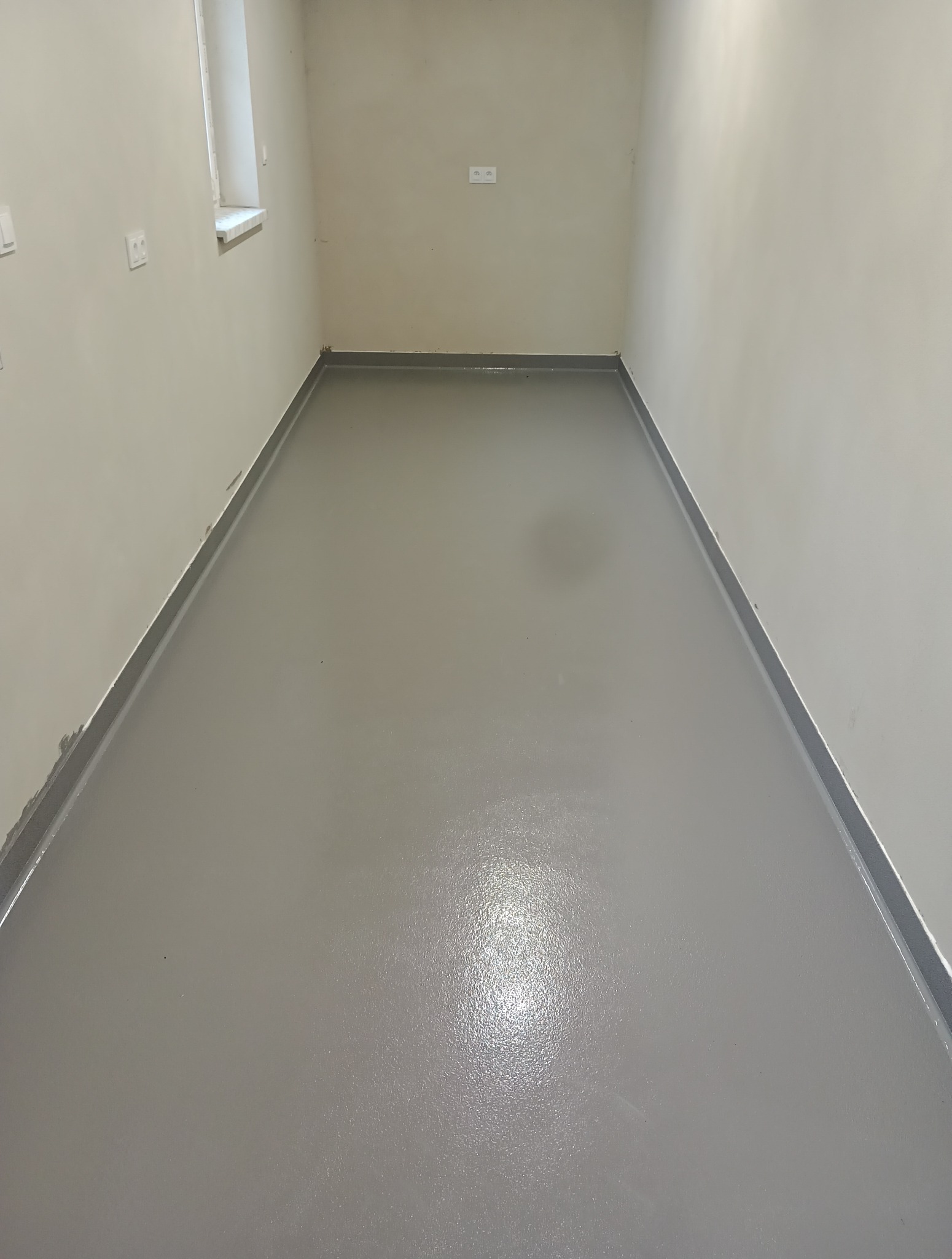 Szara, gładka posadzka żywiczna w pustym pomieszczeniu z jasnymi ścianami i widocznymi gniazdkami elektrycznymi. Minimalistyczne wykończenie podłogi.
