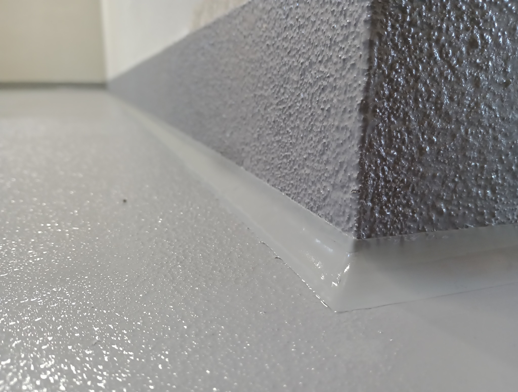 Jasnoszara, gładka posadzka żywiczna w rogu pomieszczenia, z widocznym połączeniem ściany i podłogi. Delikatna faktura ściany. Minimalistyczny detal wykończenia.