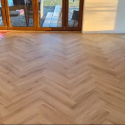 Jasne panele ułożone w jodełkę w nowym wnętrzu z dużymi oknami. Częściowo widoczne materiały montażowe, co sugeruje świeżo zakończony etap wykończenia podłogi.