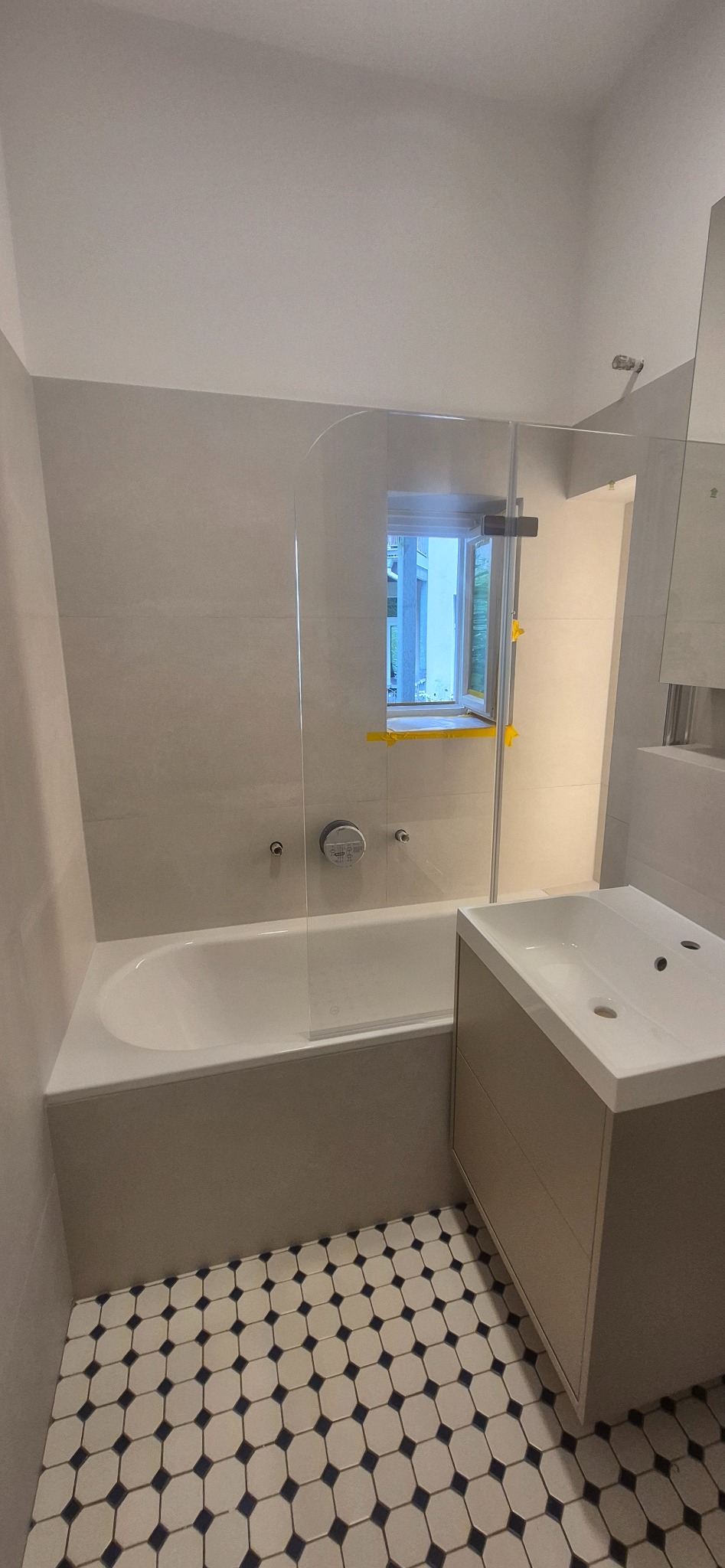 Wykończona łazienka z wanną za szklaną ścianką i umywalką z szafką. Na podłodze płytki w biało-granatową mozaikę. Okno z widokiem na zieleń.