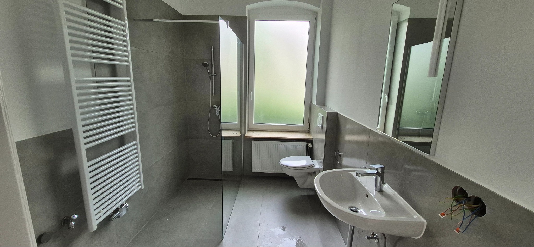 Nowoczesna łazienka z szarą glazurą, kabiną prysznicową typu walk-in, grzejnikiem drabinkowym i umywalką. Widoczne wystające przewody elektryczne.