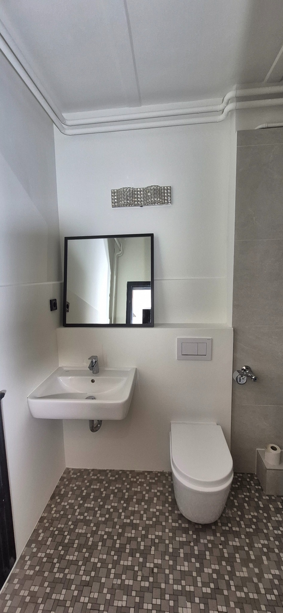 Wykończona łazienka w jasnych barwach z mozaikową podłogą. Widoczna umywalka, lustro w czarnej ramie, toaleta i grzejnik.