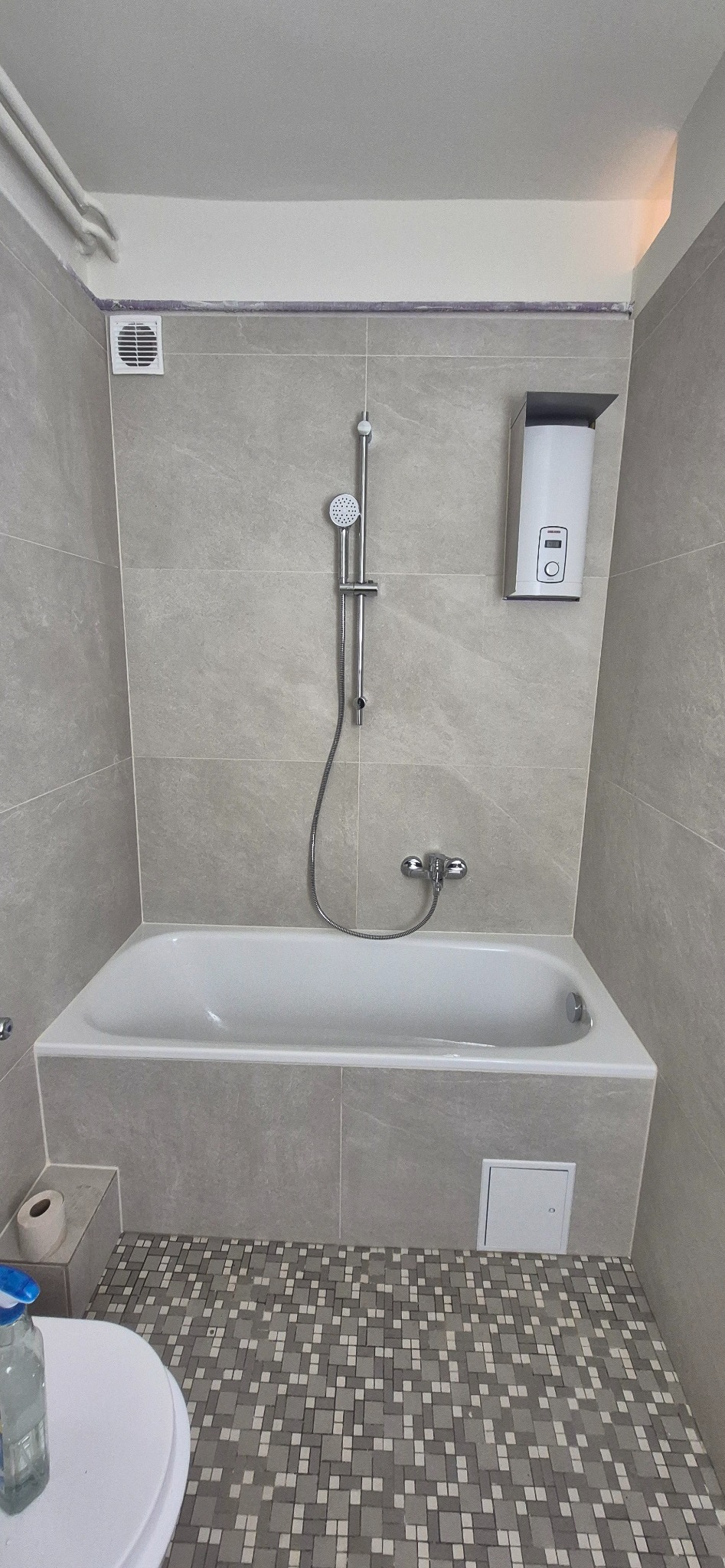 Wykończona łazienka w szarościach: płytki imitujące kamień na ścianach, mozaika na podłodze. Biała wanna, chromowana armatura i podgrzewacz wody. Minimalistyczny design.