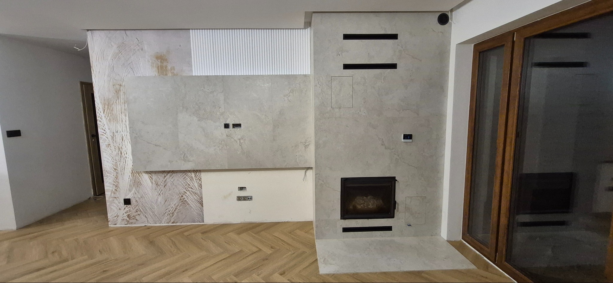 Wykończona ściana z płytami imitującymi kamień, z wbudowanym kominkiem i elementami dekoracyjnymi, na podłodze parkiet ułożony w jodełkę.