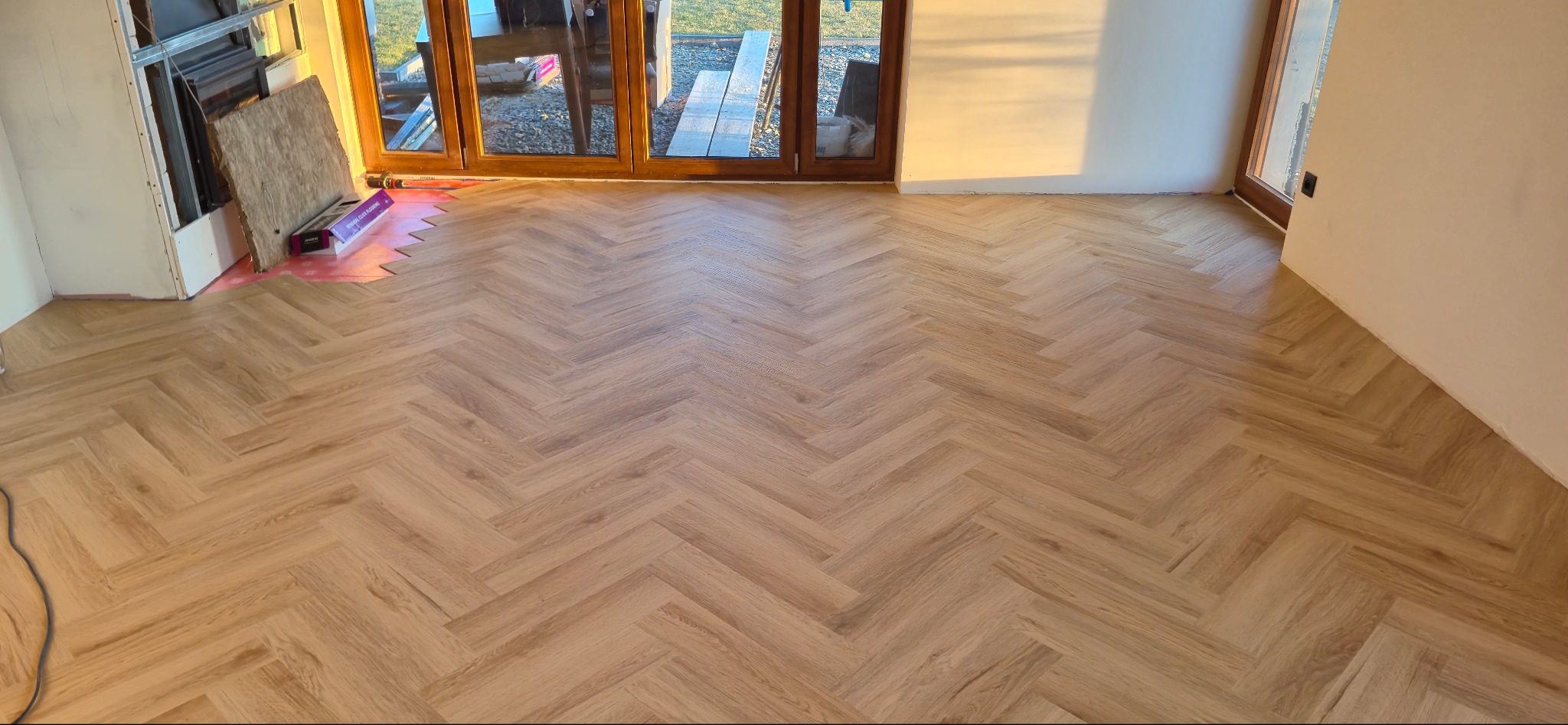 Jasne panele ułożone w jodełkę w nowym wnętrzu z dużymi oknami. Częściowo widoczne materiały montażowe, co sugeruje świeżo zakończony etap wykończenia podłogi.