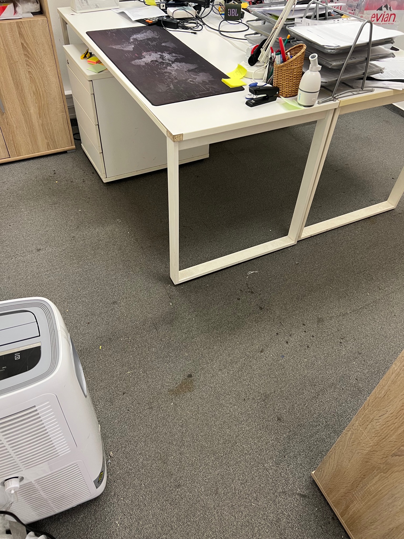 Szary dywan biurowy z plamami, białe biurko z akcesoriami, klimatyzator przenośny. Widok z góry, neutralne oświetlenie.