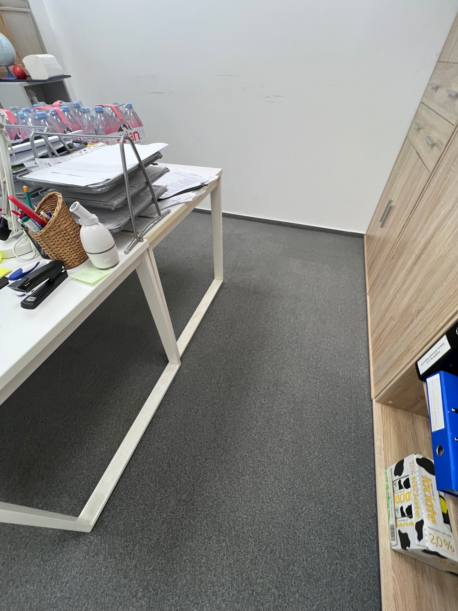 Częściowy widok biura: białe biurko z akcesoriami (zszywacz, długopisy, nawilżacz powietrza), szare wykładziny i drewniana szafa w tle. Ujęcie z bliska.