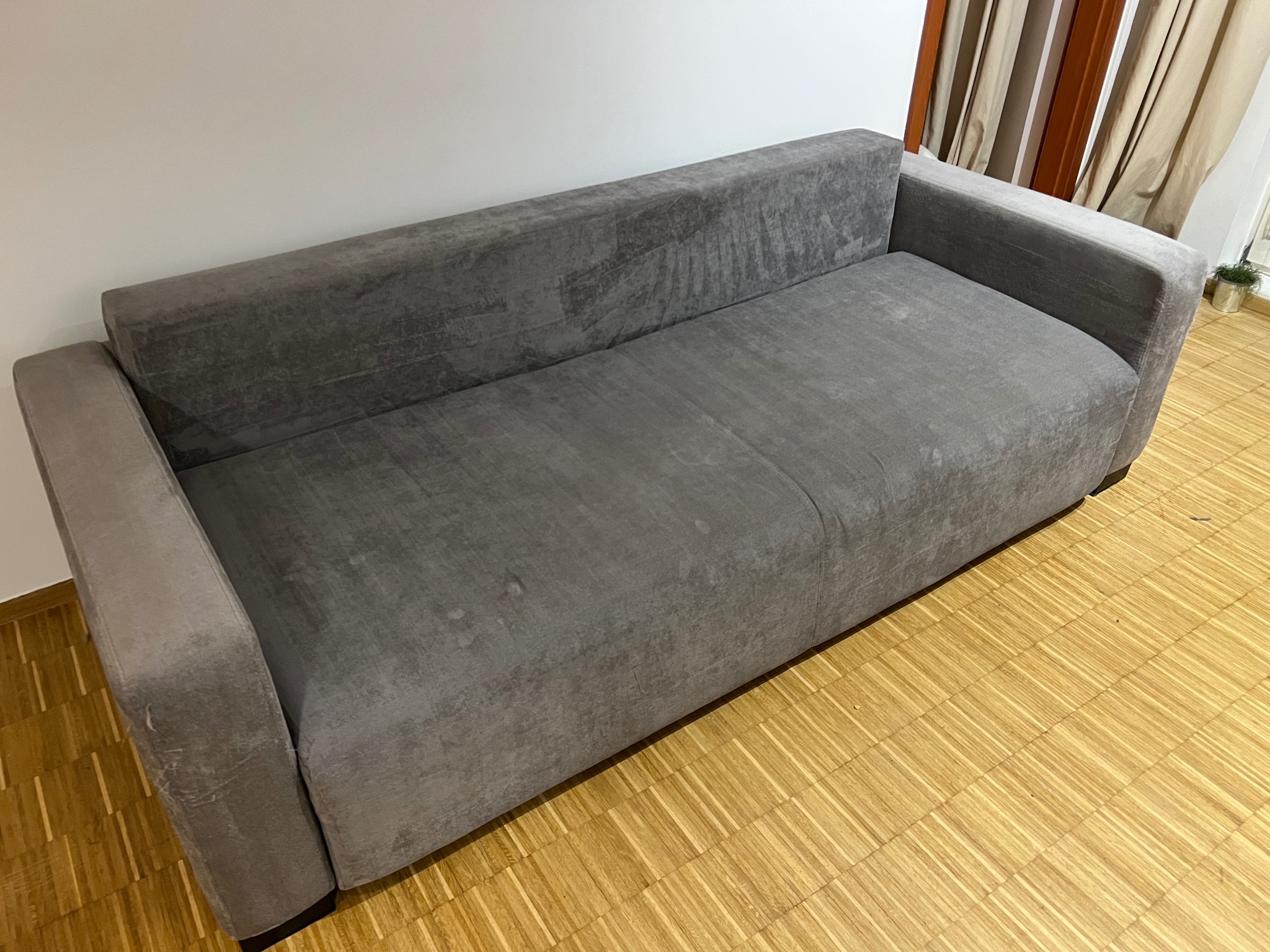 Szara, welurowa kanapa na parkiecie, z widocznymi śladami użytkowania na boku, sugerująca potrzebę odświeżenia tapicerki. Neutralne tło, stonowane światło.