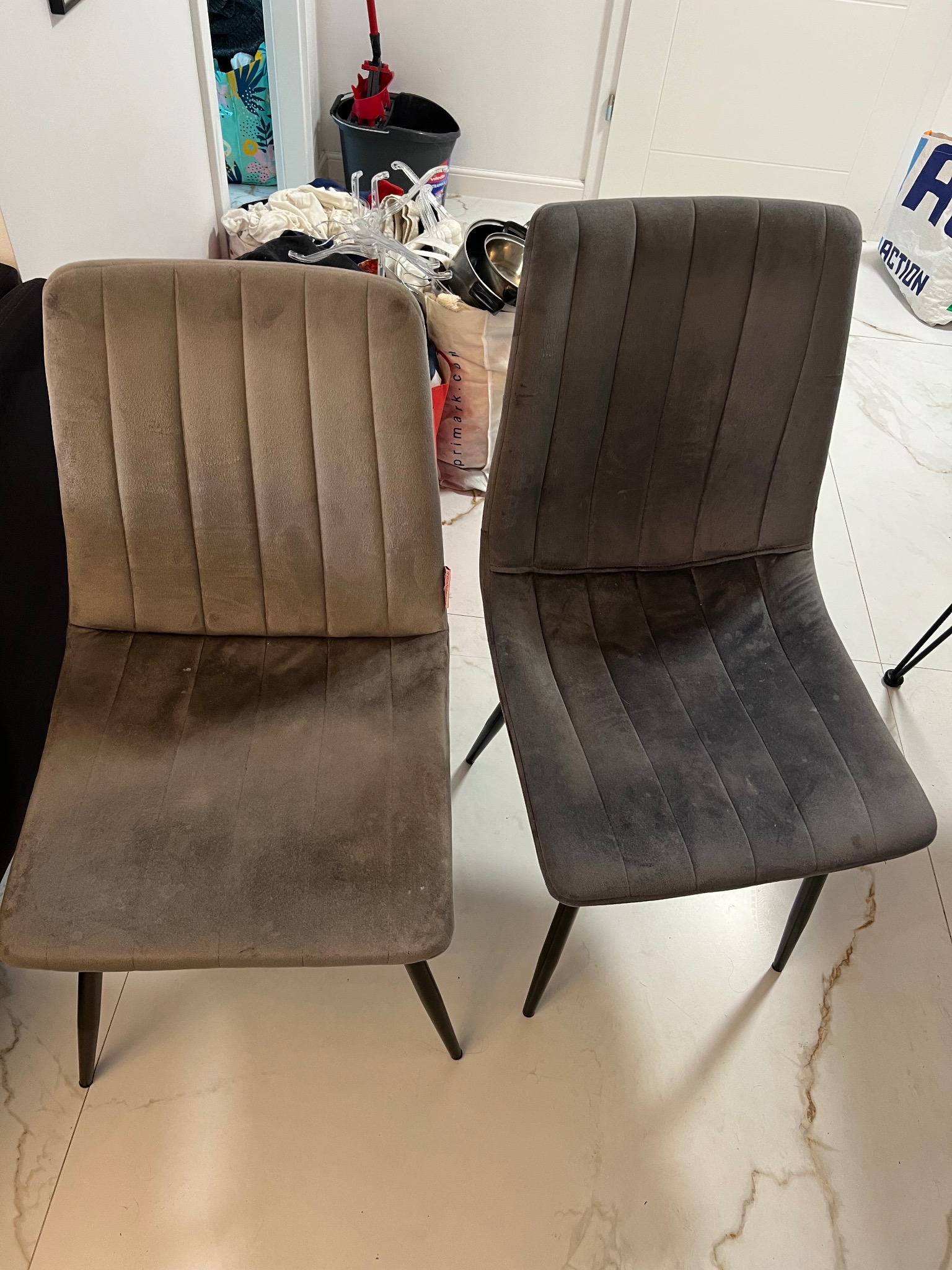 Dwa welurowe krzesła (beżowe i szare) z zabrudzeniami na siedziskach, stojące na marmurowej podłodze. W tle widoczne akcesoria do sprzątania i torba.
