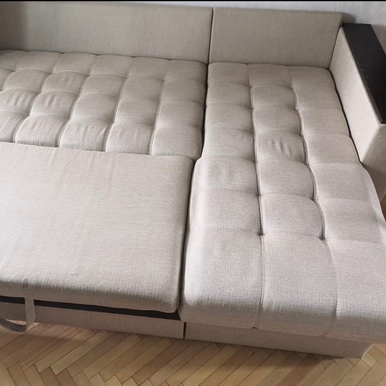 Jasna, rozłożona kanapa z pikowanymi siedziskami i drewnianym podłokietnikiem, ustawiona na parkiecie. Widoczny wysunięty element do spania.