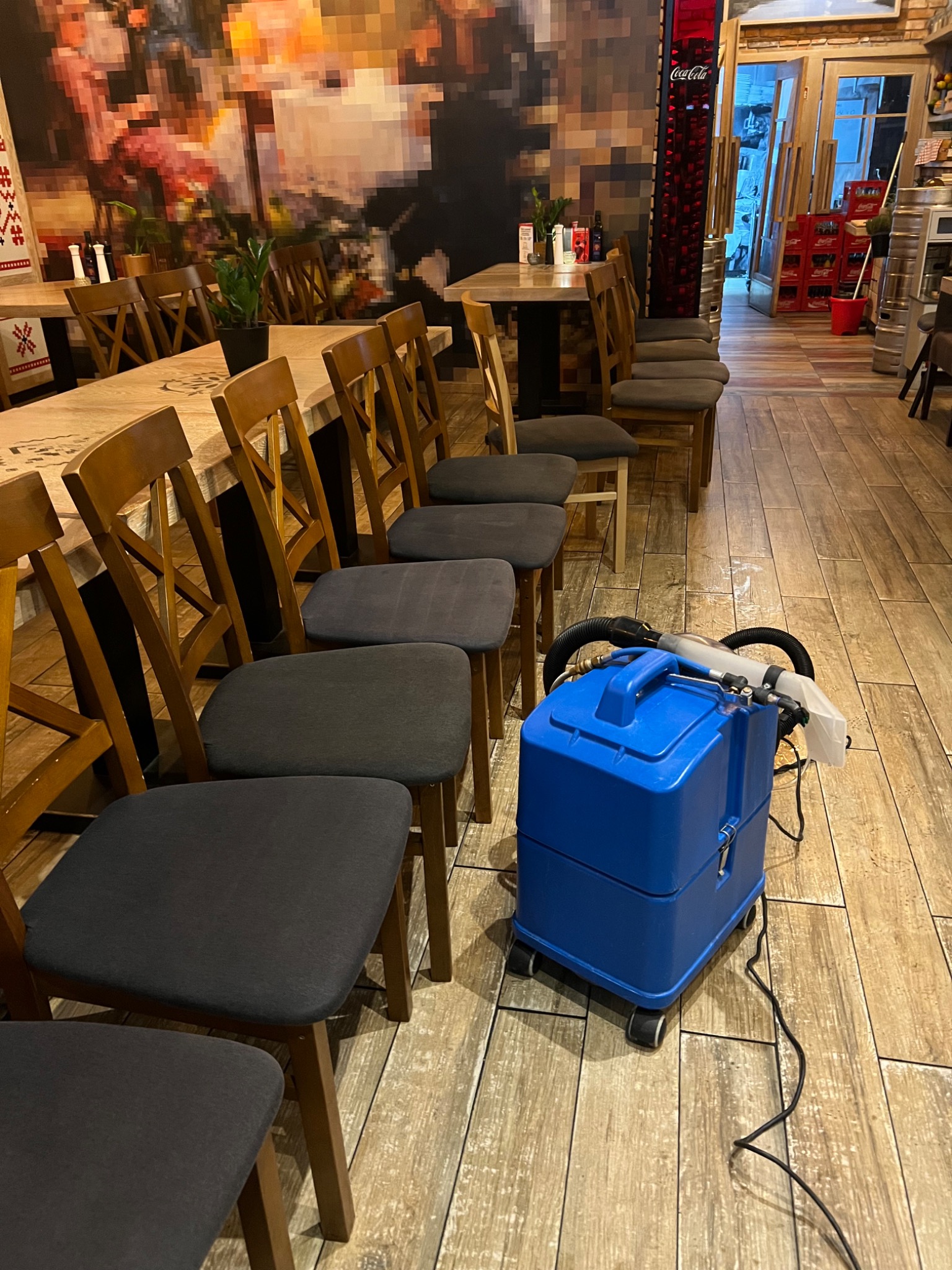 Niebieski odkurzacz piorący stoi na drewnianej podłodze w restauracji, gotowy do prania tapicerki krzeseł. W tle stoły i krzesła, dekoracje na ścianie.