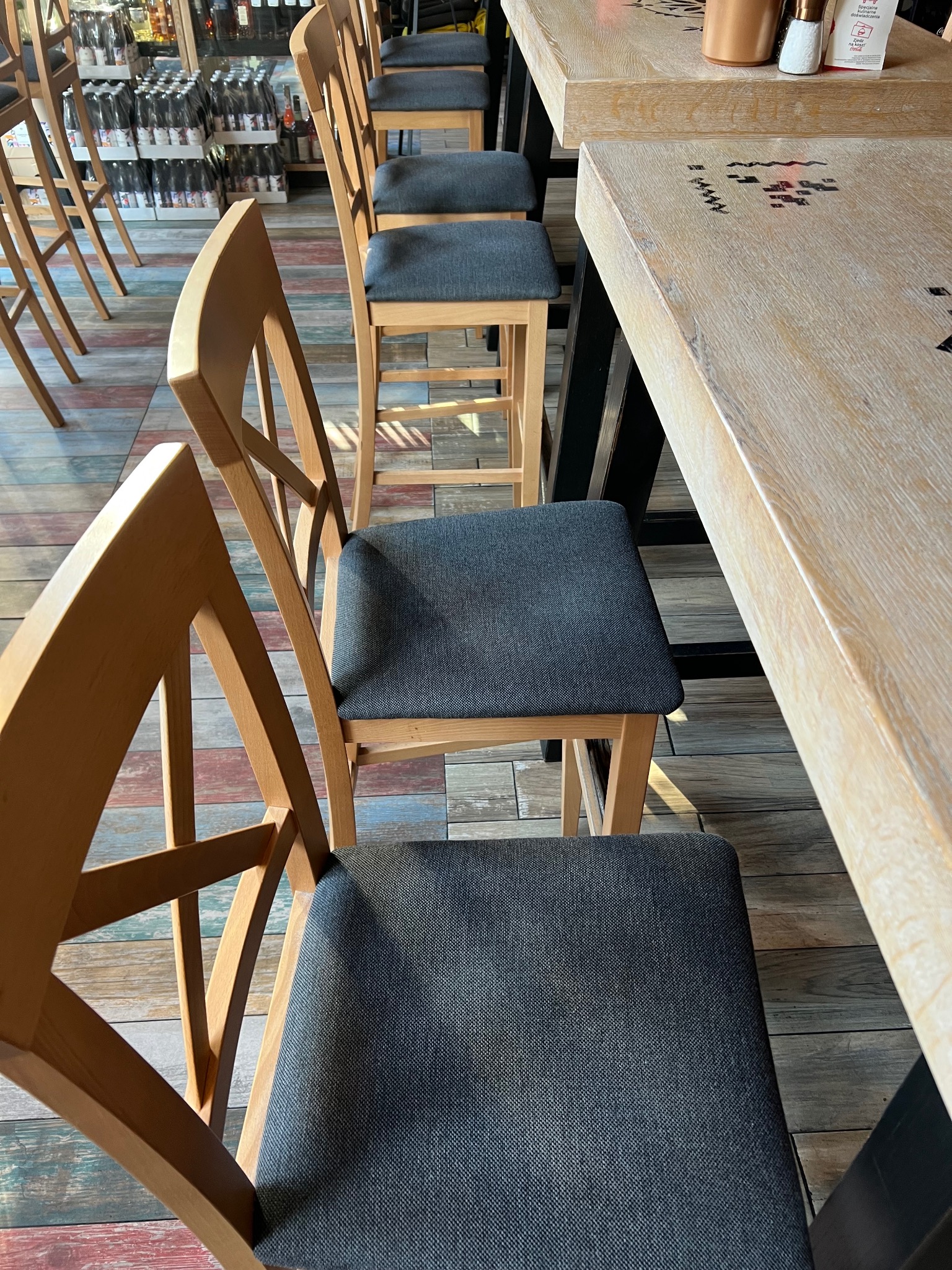 Rząd drewnianych krzeseł barowych z szarymi siedziskami ustawionych przy jasnym, drewnianym blacie. W tle widoczne butelki na półkach i podłoga z kolorowych desek.