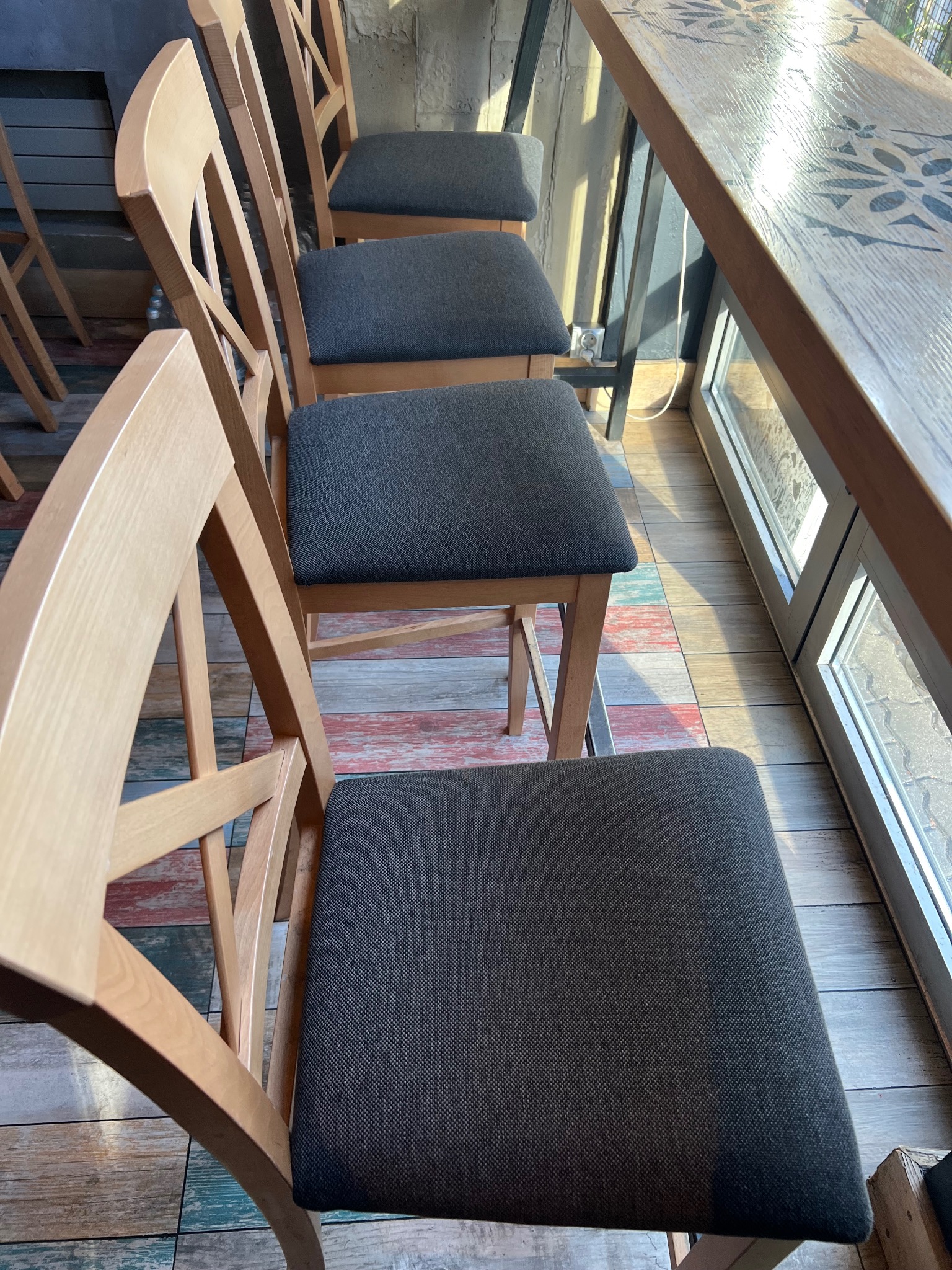 Rząd drewnianych krzeseł z szarymi siedziskami przy stole z dekoracyjnym wzorem, ustawionych na podłodze z kolorowych desek. Widok z góry, blisko okna.