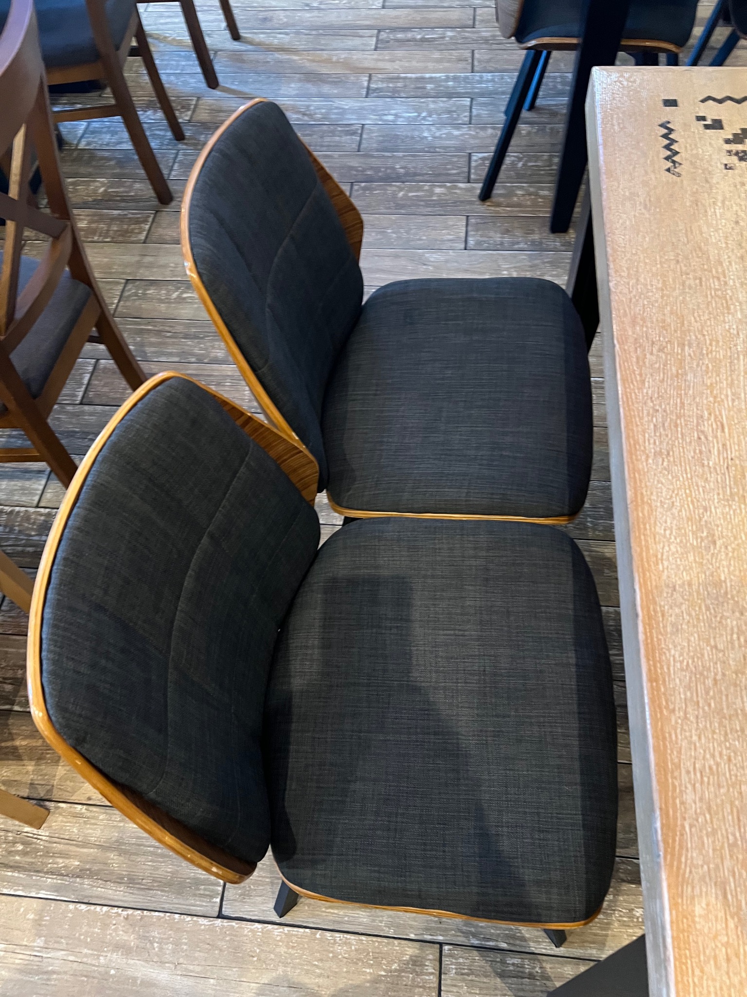 Dwa krzesła tapicerowane ciemnoszarą tkaniną, ustawione przy jasnym, drewnianym stole. Ujęcie z góry, widoczne wzory na blacie i drewniana podłoga.