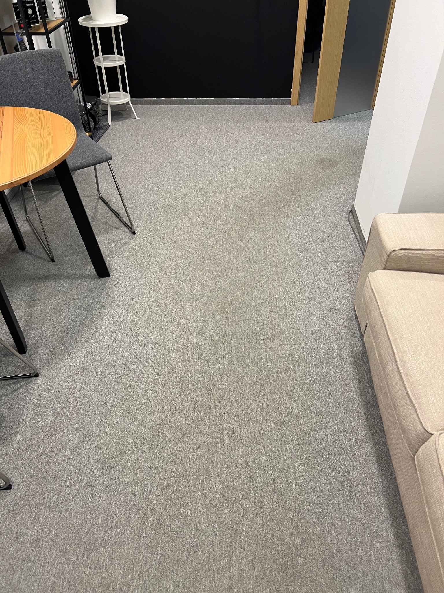 Wnętrze biura z szarym dywanem, stołem z drewnianym blatem i krzesłem. Widoczny fragment sofy w jasnym odcieniu. Minimalistyczny wystrój, czarna ściana w tle.