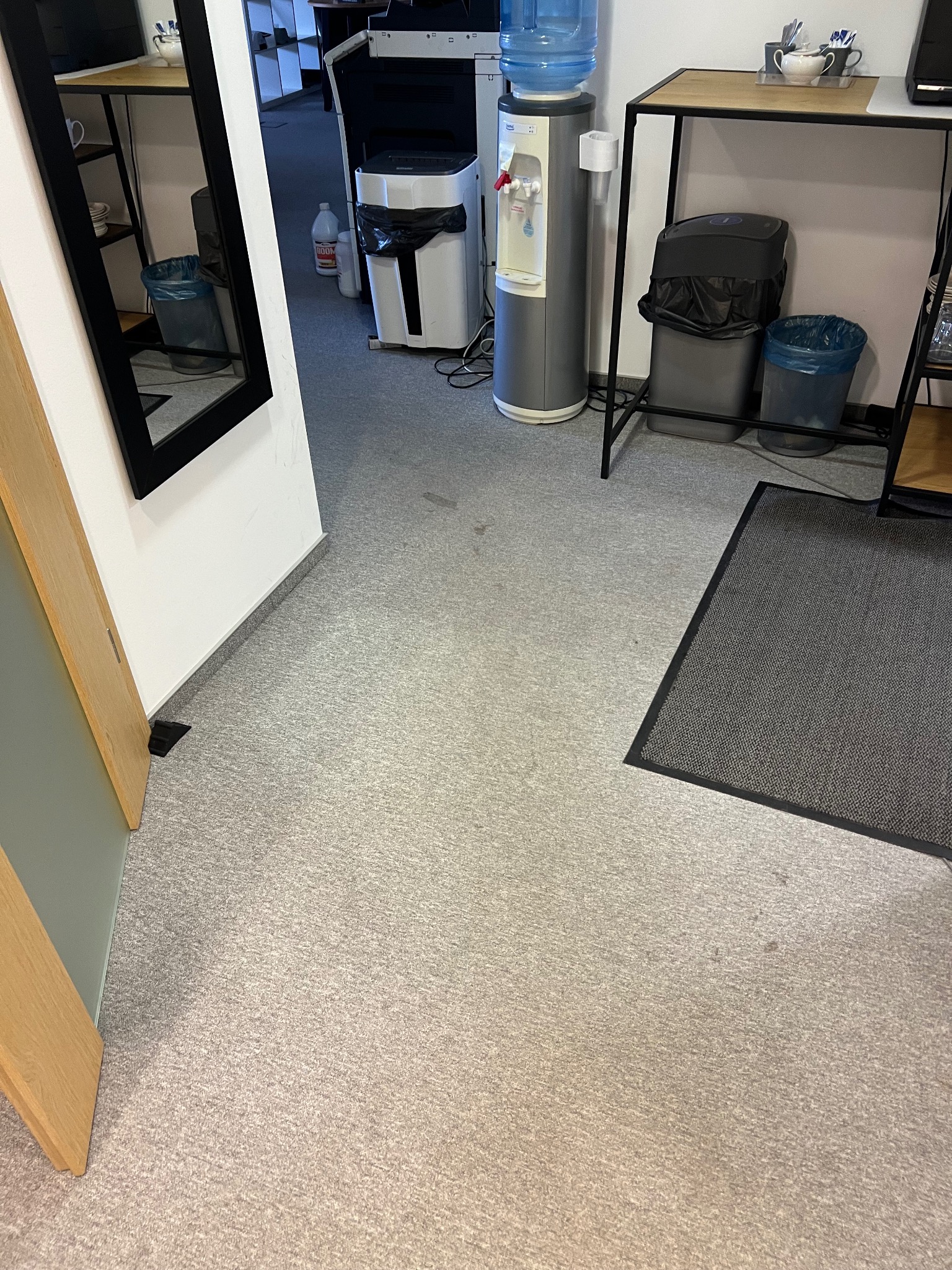 Jasna wykładzina biurowa z ciemnymi plamami, widoczny fragment dywanu, kosze na śmieci i dystrybutor wody. W tle biurowe wyposażenie.