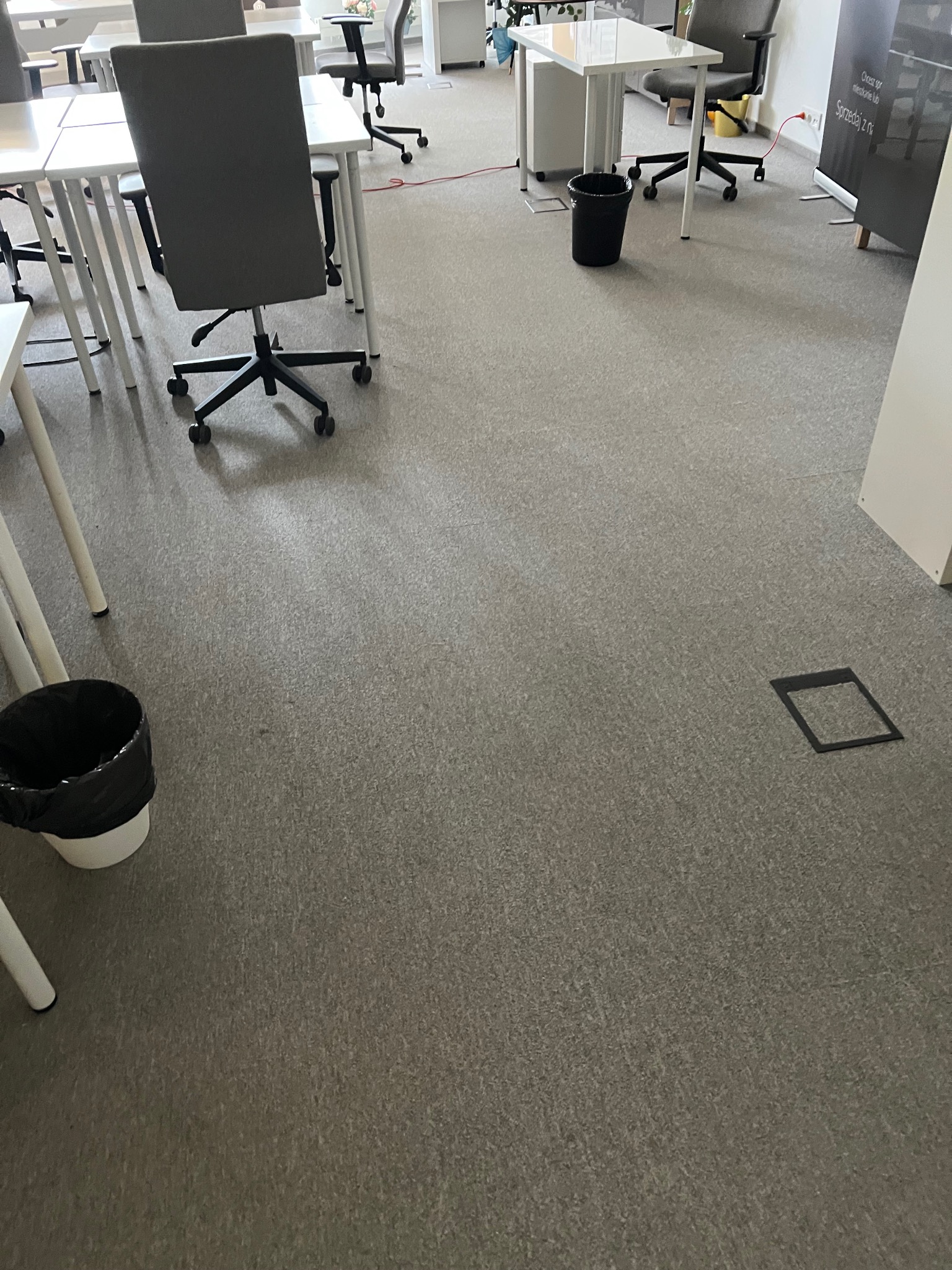 Wnętrze biura z szarym dywanem, białymi biurkami i krzesłami biurowymi. Widoczny kosz na śmieci i gniazdo podłogowe. Jasne oświetlenie.