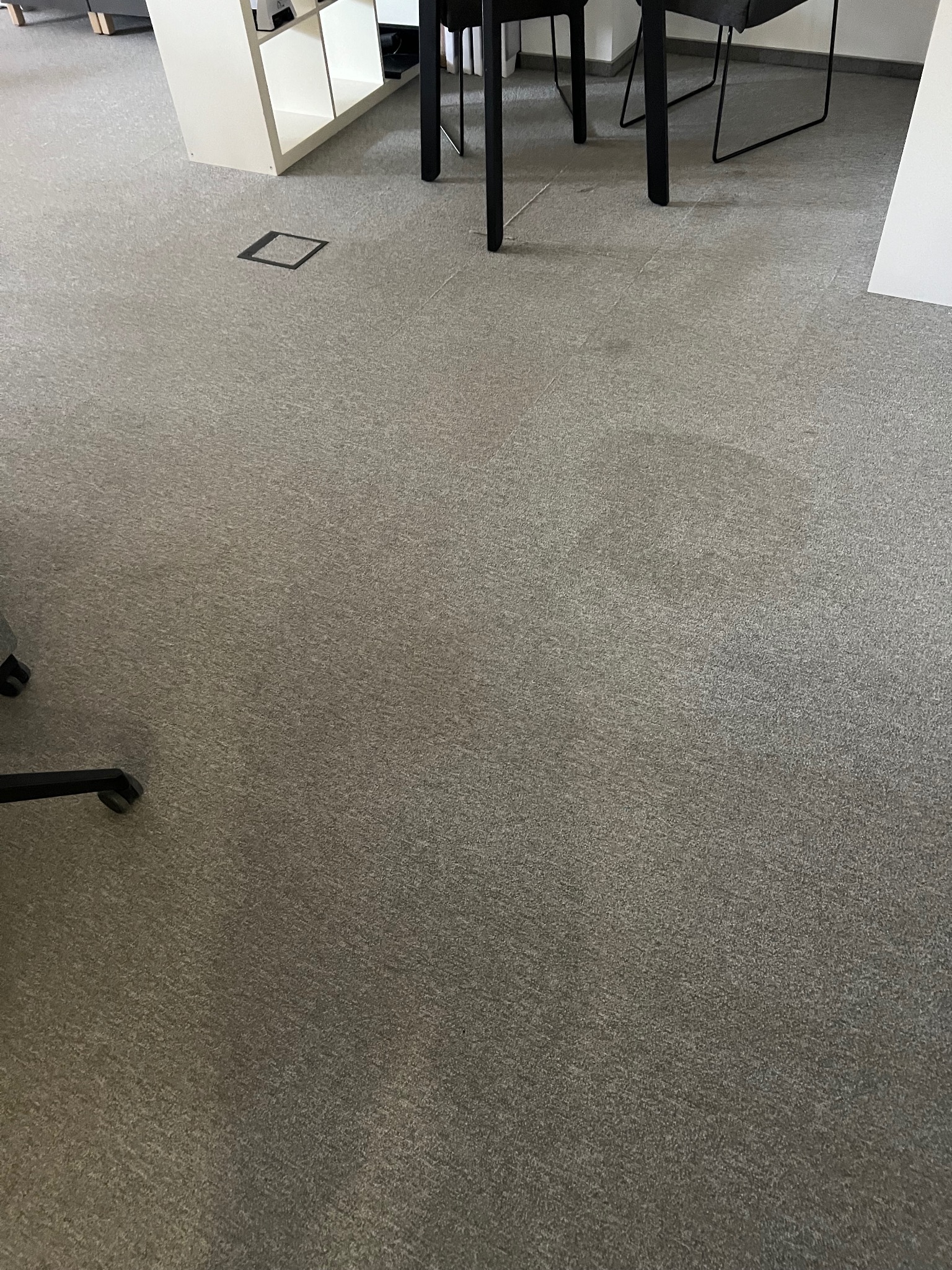 Szary dywan biurowy z ciemniejszą plamą, widoczne nogi krzeseł i fragment regału z białymi półkami w tle. Tekstura dywanu jest wyraźna.