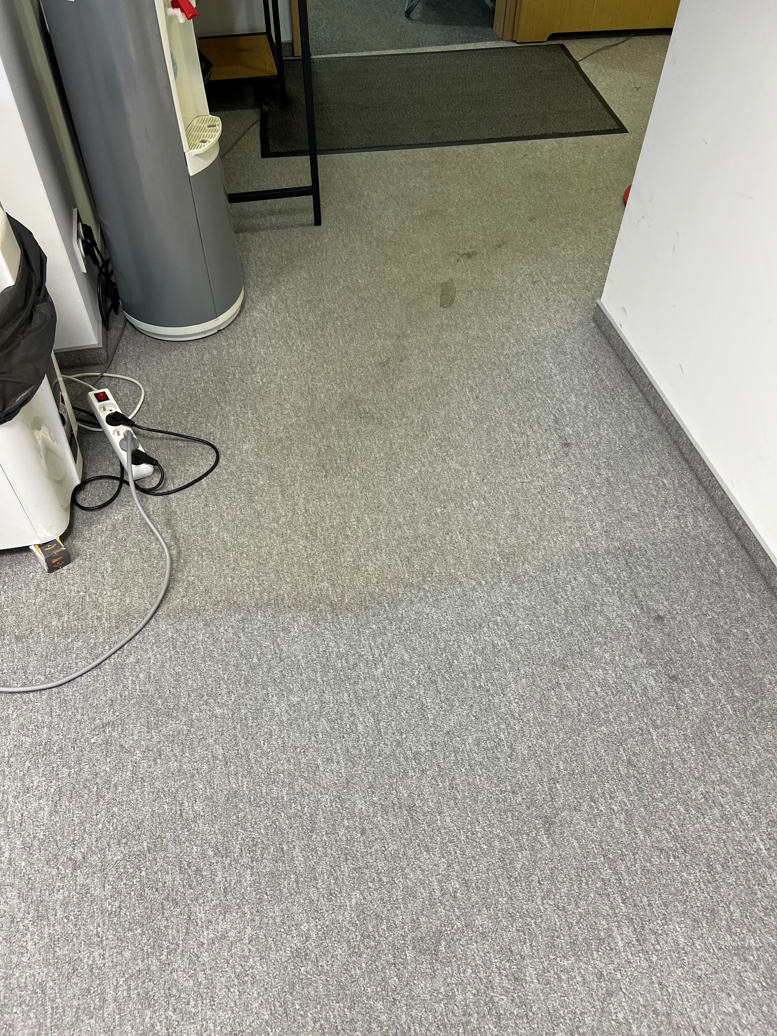 Szara wykładzina biurowa z widocznymi plamami, obok ściana i dystrybutor wody. Widoczne ślady użytkowania i zabrudzenia na powierzchni dywanu.