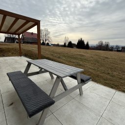 HolzWood Michał Bednarczyk - Drewniany stół piknikowy z szarymi poduszkami na tarasie z widokiem na krajobraz. W tle pergola z zadaszeniem i domy. Pochmurne niebo.