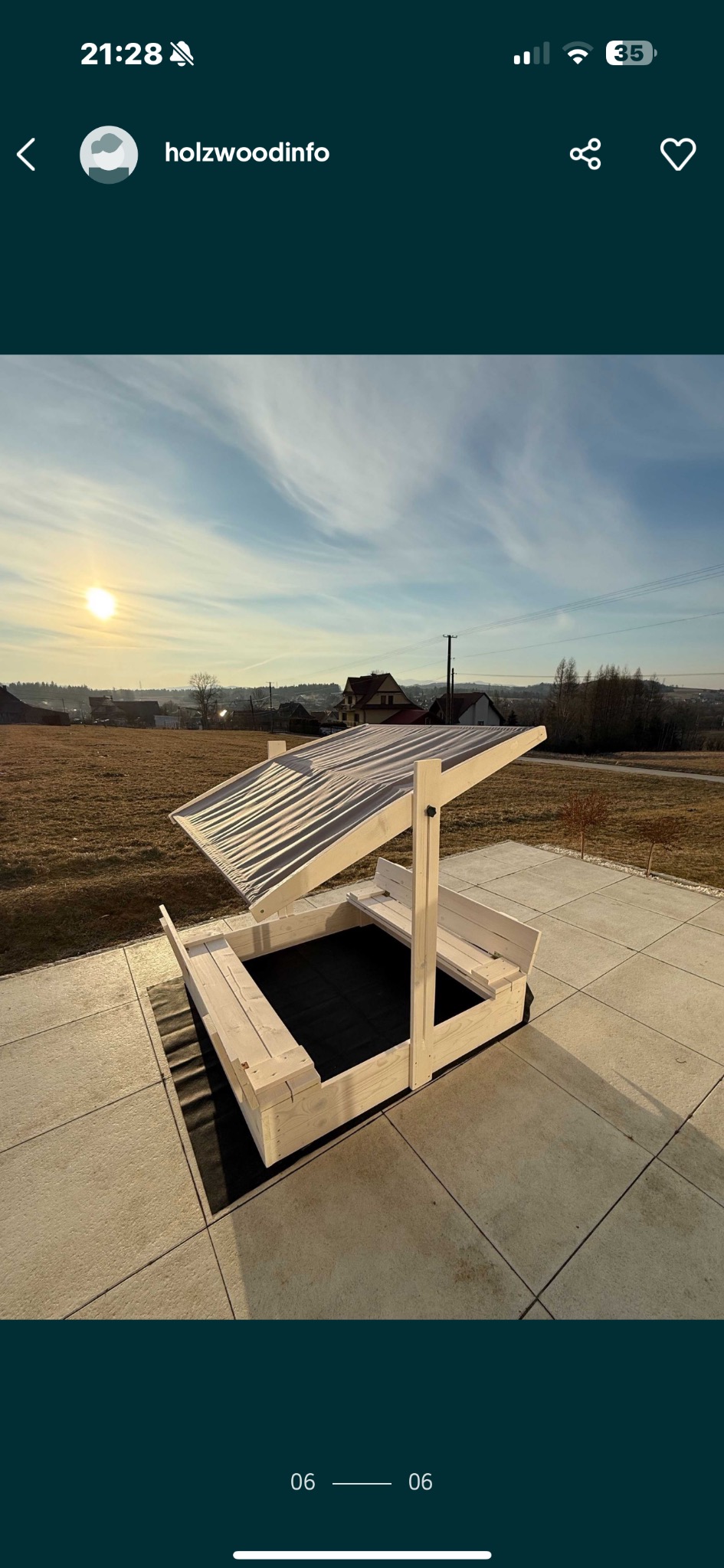 Drewniana piaskownica z zadaszeniem i siedziskami, ustawiona na betonowej kostce, w tle krajobraz wiejski w słoneczny dzień.