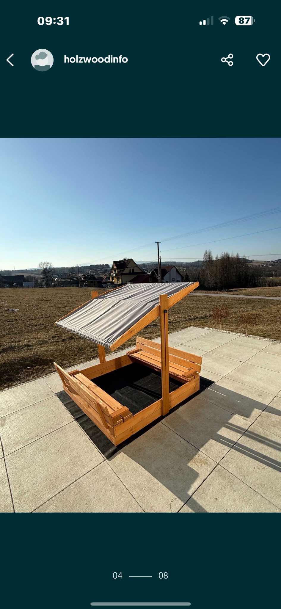 Drewniana piaskownica z zadaszeniem na tarasie z jasnych płyt, w tle zabudowa jednorodzinna i błękitne niebo. Widok z góry.